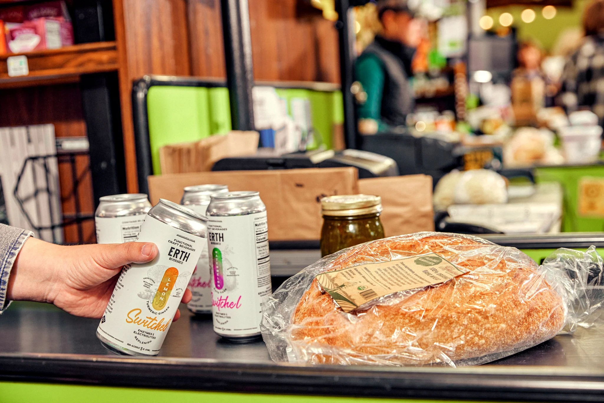 Hand holding three cans of functional beverage called Earth with ginger, lemon, turmeric, and honey flavors. In the background, there is a supermarket or store setting with shelves of various products, a person wearing a vest and green shirt, and a counter with packaged food items, including a piece of salmon in plastic wrap.