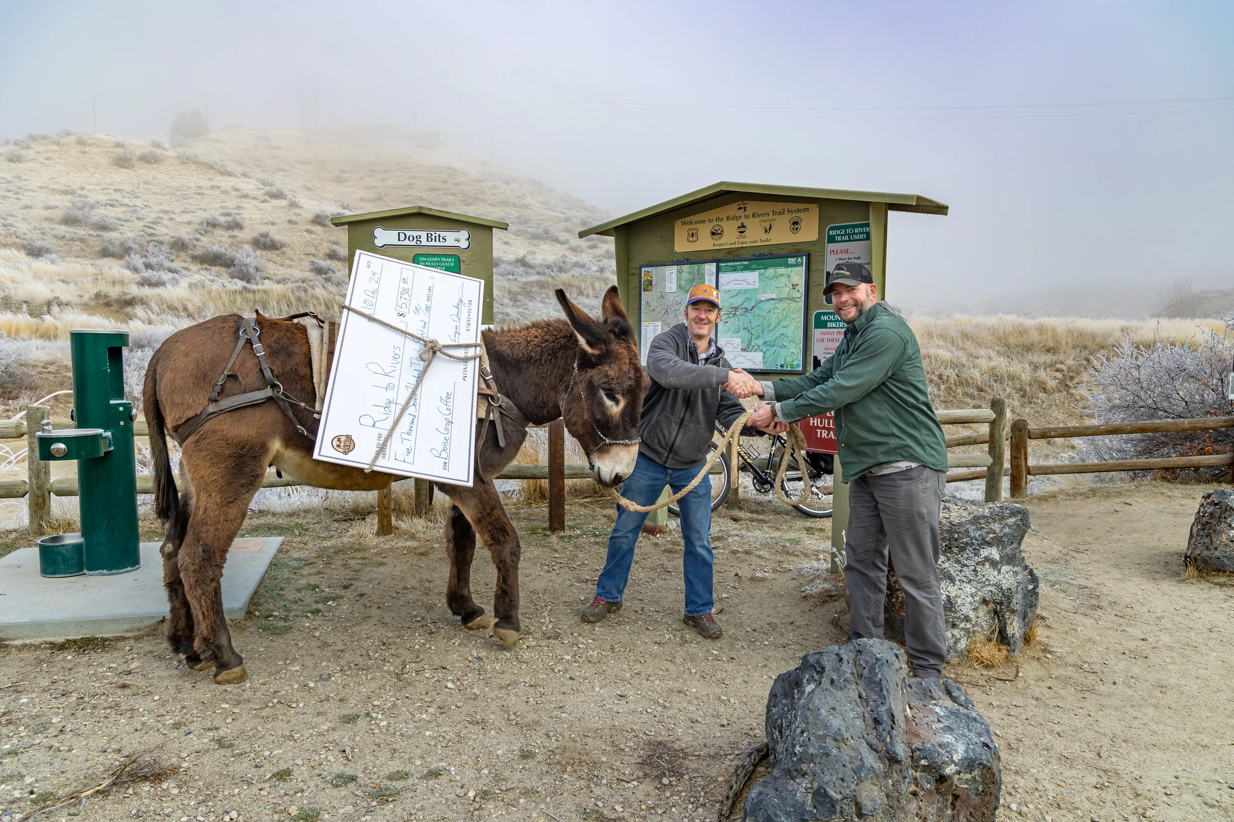 Two men shake hands next to a donkey at a trailhead. The donkey is carrying a load and a sign for dog bites. The background shows a foggy hillside and a posted map of the trail system.