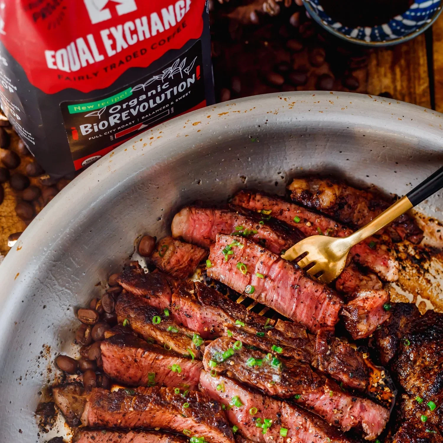 Sliced cooked steak garnished with chopped green onions in a metal pan. Behind the pan is a package of organic biorevolution coffee and a bowl of dark roasted coffee beans.