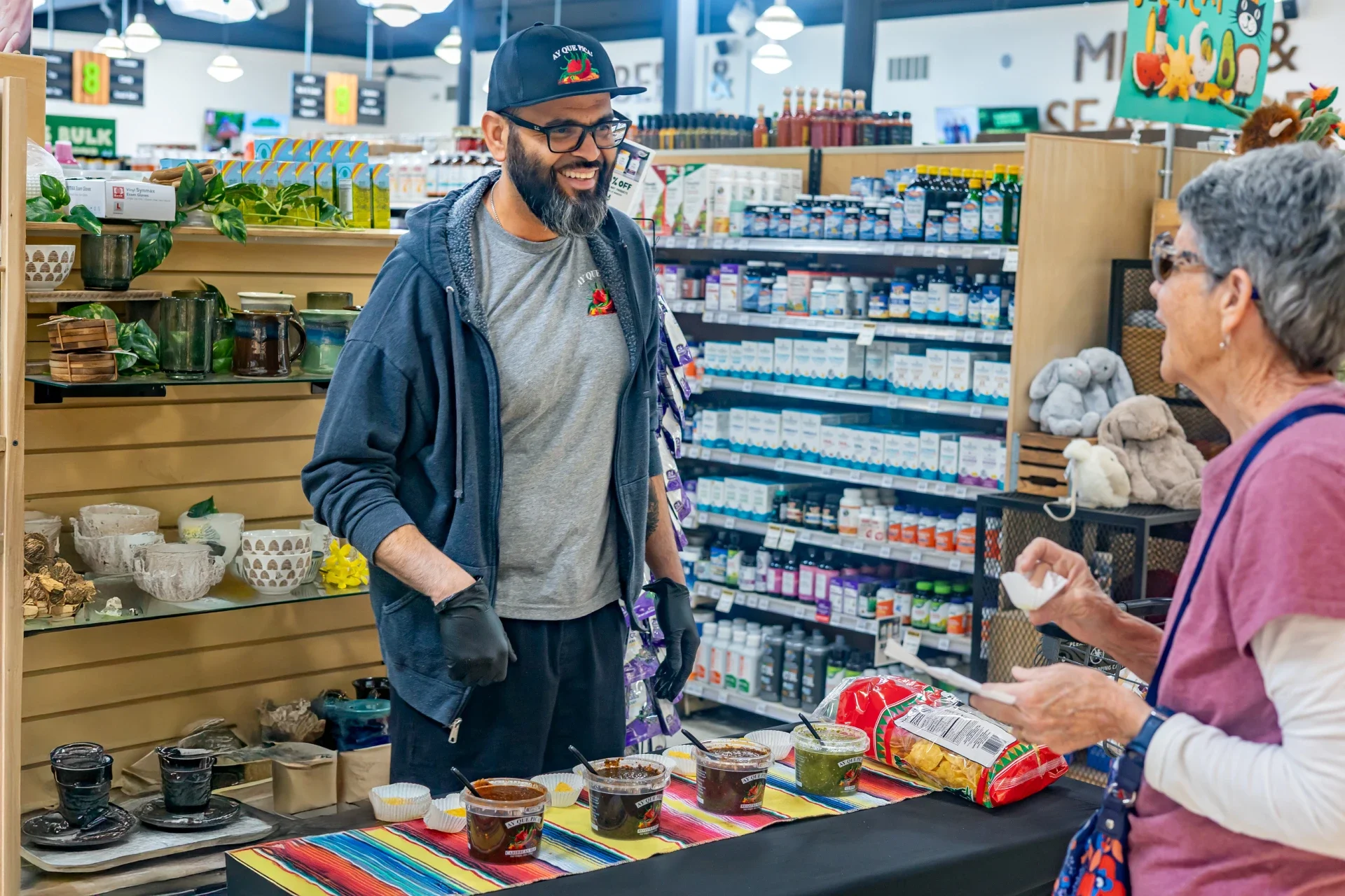 Man with glasses, beard, and dark clothing smiling and talking to an older woman with gray hair and sunglasses at a food sampling stand inside a store.
