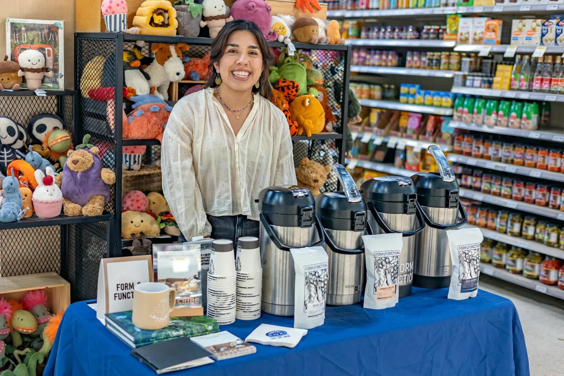 Woman smiling behind a table with coffee brewers and cups in a store aisle, with shelves of snacks and plush toys behind her
