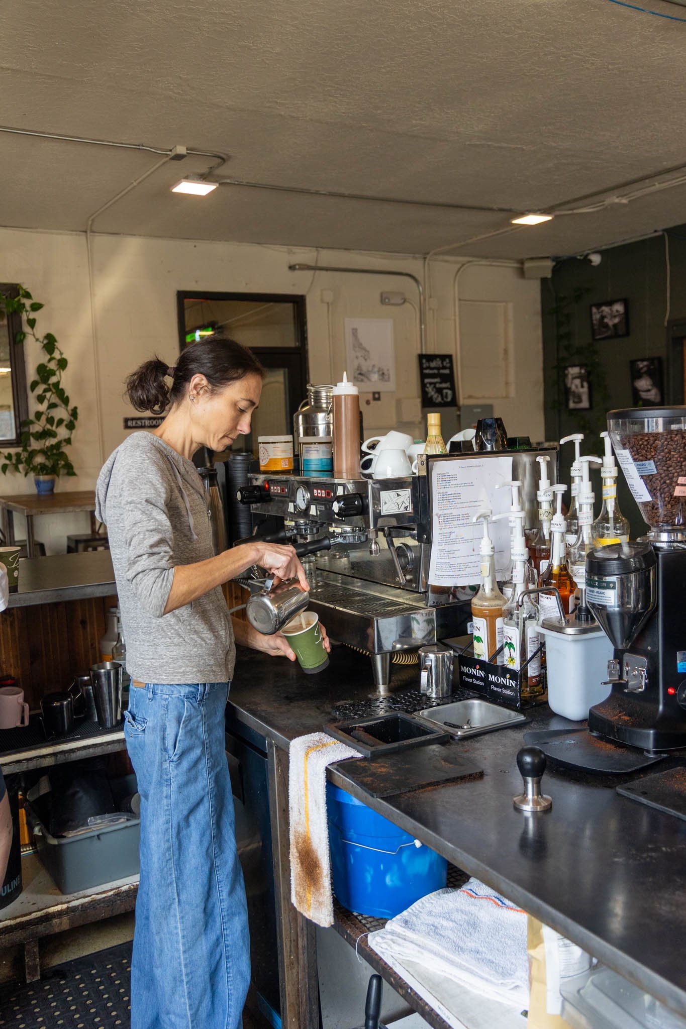 A woman making coffee behind a coffee bar with a latte art pitcher and espresso machine, in a cozy cafe with framed pictures and a plant on the wall.