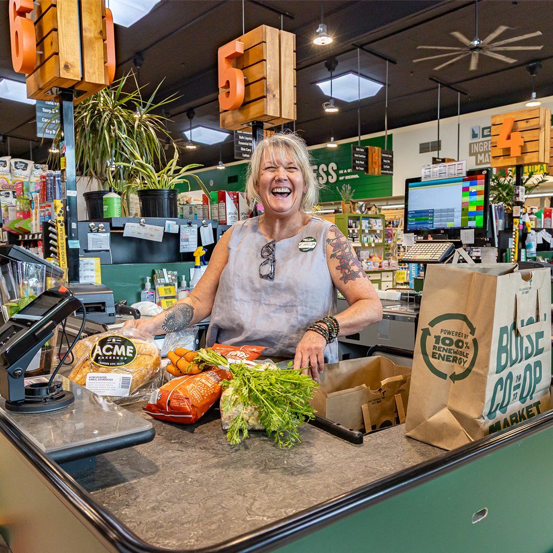 A cheerful woman with tattoos and blonde hair at a grocery checkout counter, surrounded by fresh vegetables and grocery bags in a store with signs and shelves in the background.