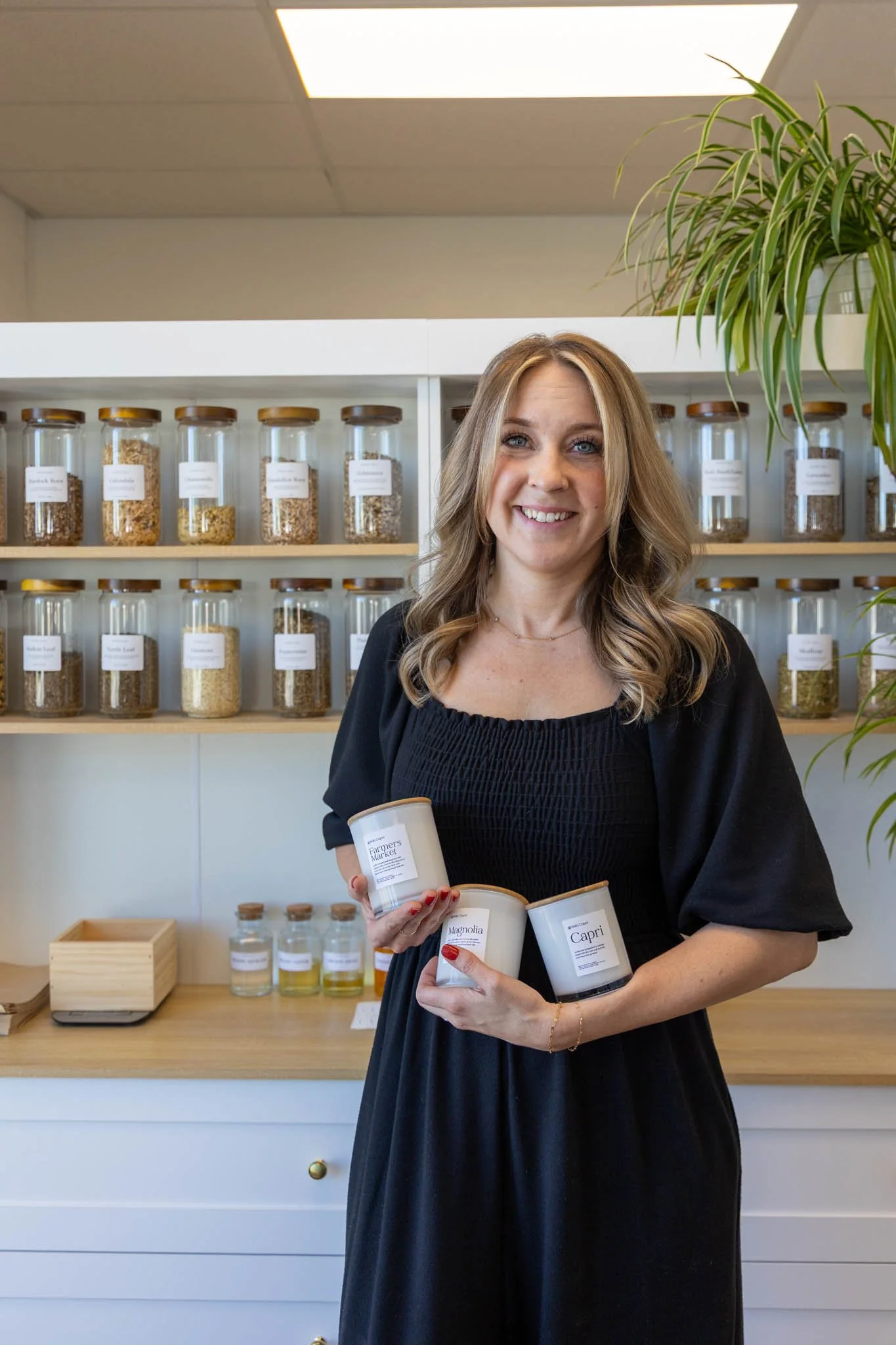 Woman with wavy hair and a black dress holding candles with labels, standing in front of shelves filled with glass jars of grains and dry ingredients in a modern workspace.
