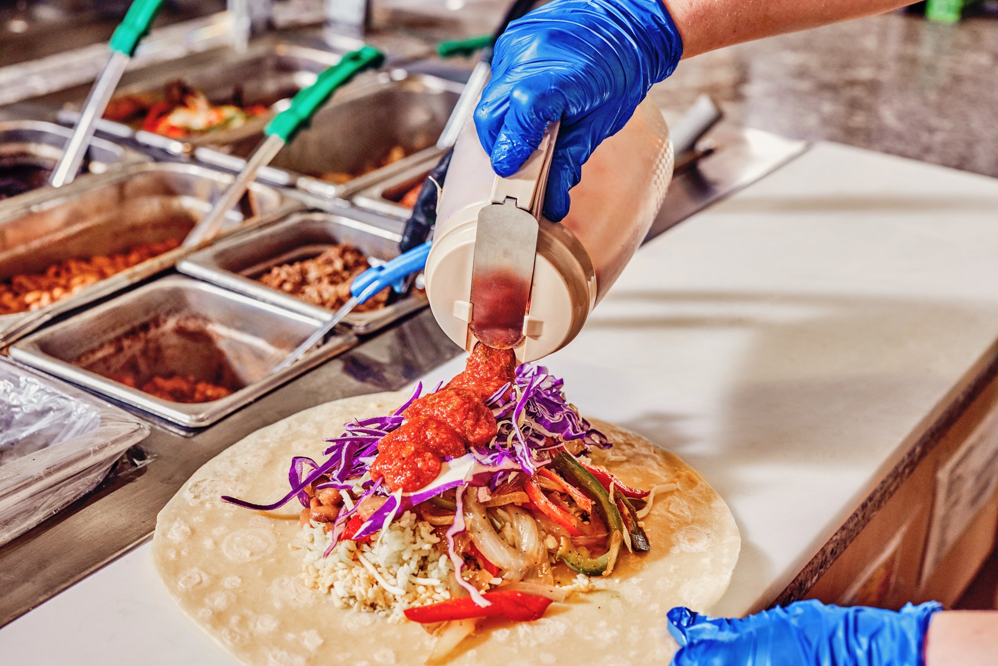 A person wearing blue gloves is filling a taco shell with seasoned meat, shredded vegetables, and rice from a variety of containers, with many sauce containers in the background.