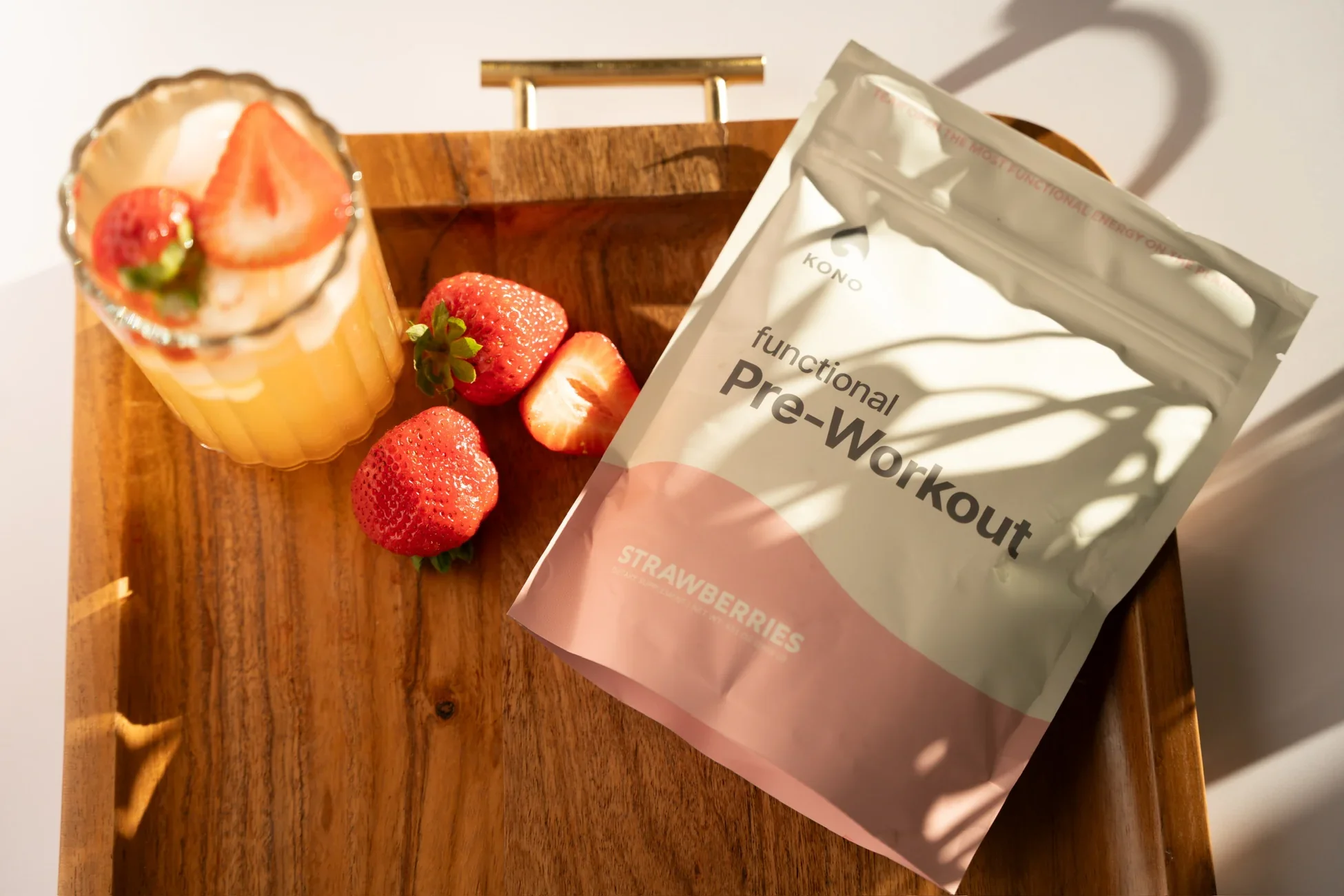 A wooden table holds a glass of strawberry smoothie topped with sliced strawberries, whole strawberries, a packet of Kono functional pre-workout, and a slice of strawberry.