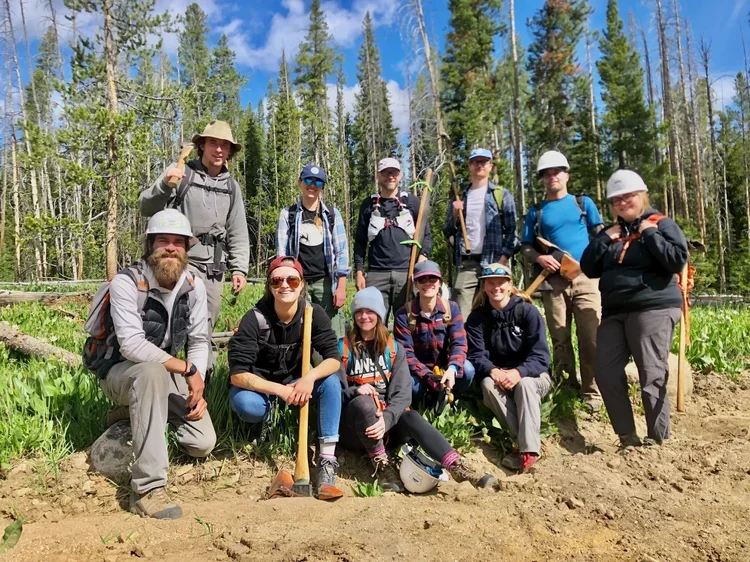Group of people in outdoor gear posing for a photo in a forested area, some holding shovels and wearing helmets.