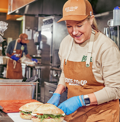 A woman wearing a brown cap and apron, smiling while preparing sandwiches in a commercial kitchen.