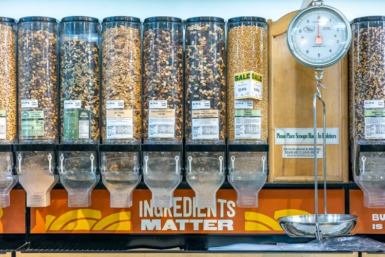 Bulk cereal dispensers filled with various cereals in a grocery store, with a balance scale and a sign that reads 'Please Place Scoops Back and in Utensils'.
