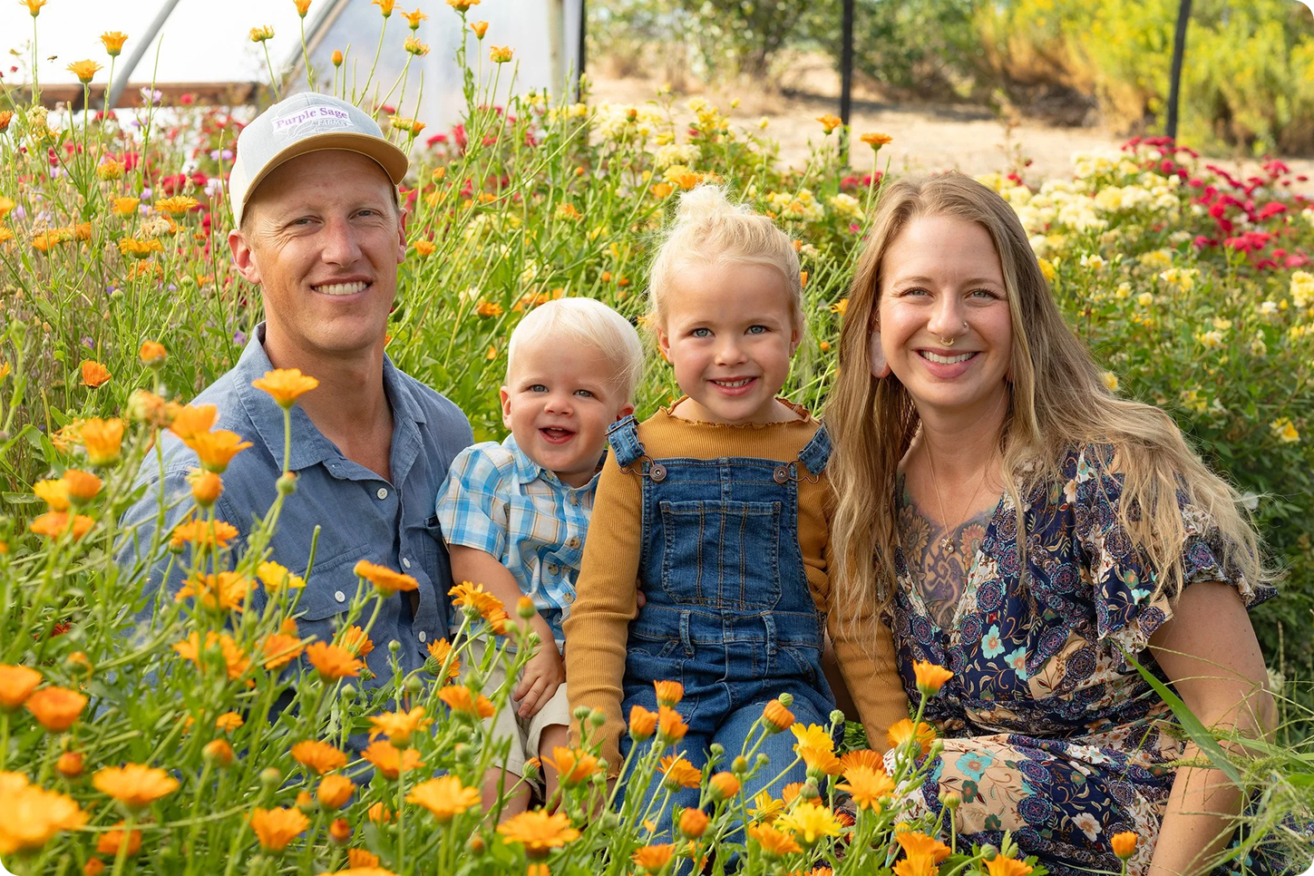 A family of four sitting among yellow and orange flowers in a garden, smiling at the camera. The father wears a cap and a blue shirt, the mother has long blonde hair and wears a patterned dress, the daughter has blonde hair in a bun and wears a mustard shirt with denim overalls, and the young boy wears a blue and white plaid shirt.