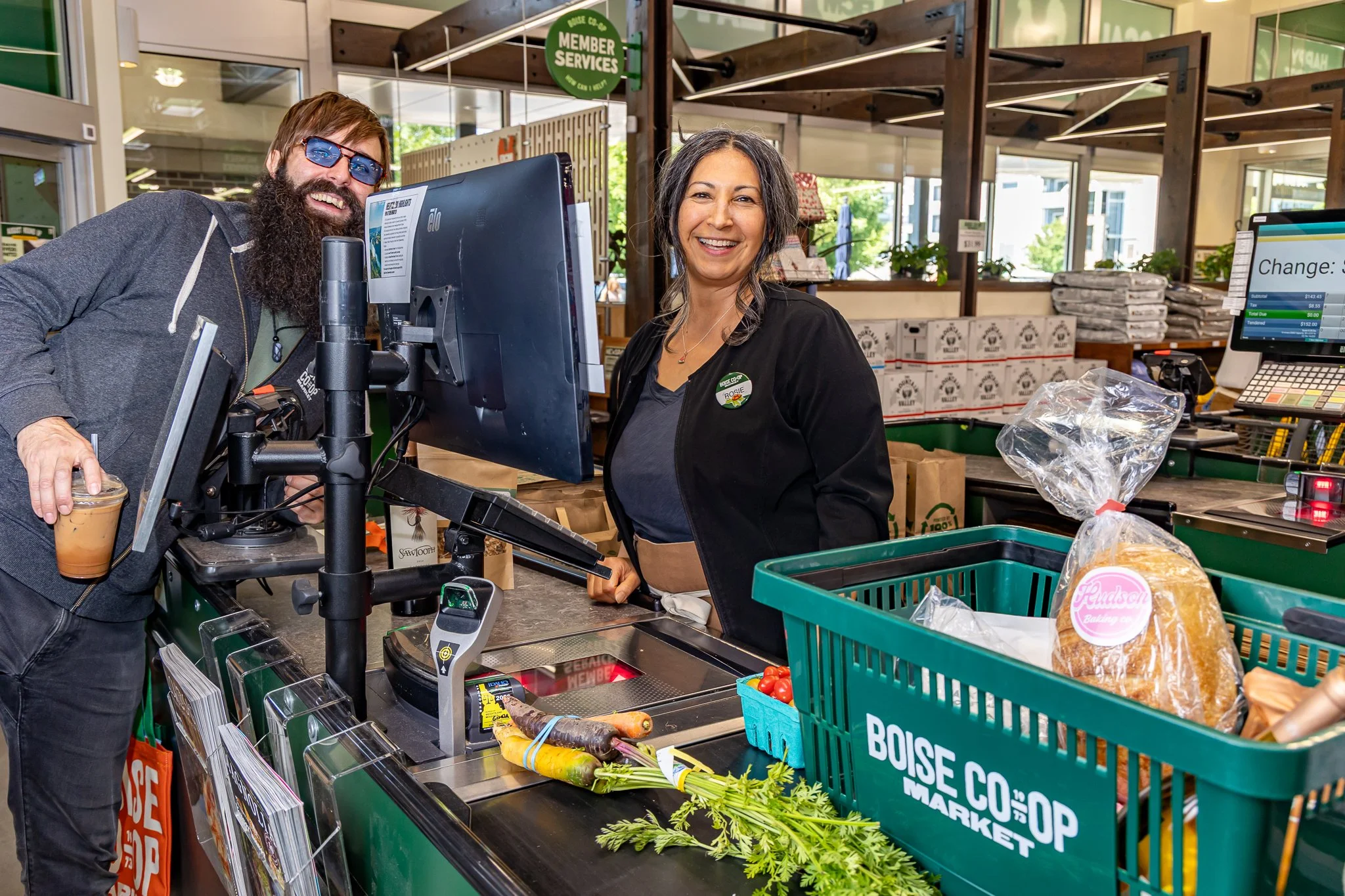 A man and woman smiling at checkout counter in grocery store, with produce and grocery bags nearby.