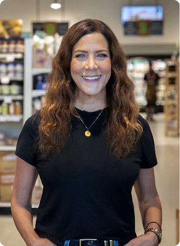 A woman with long, wavy brown hair wearing a black t-shirt and a gold necklace smiling inside a store.