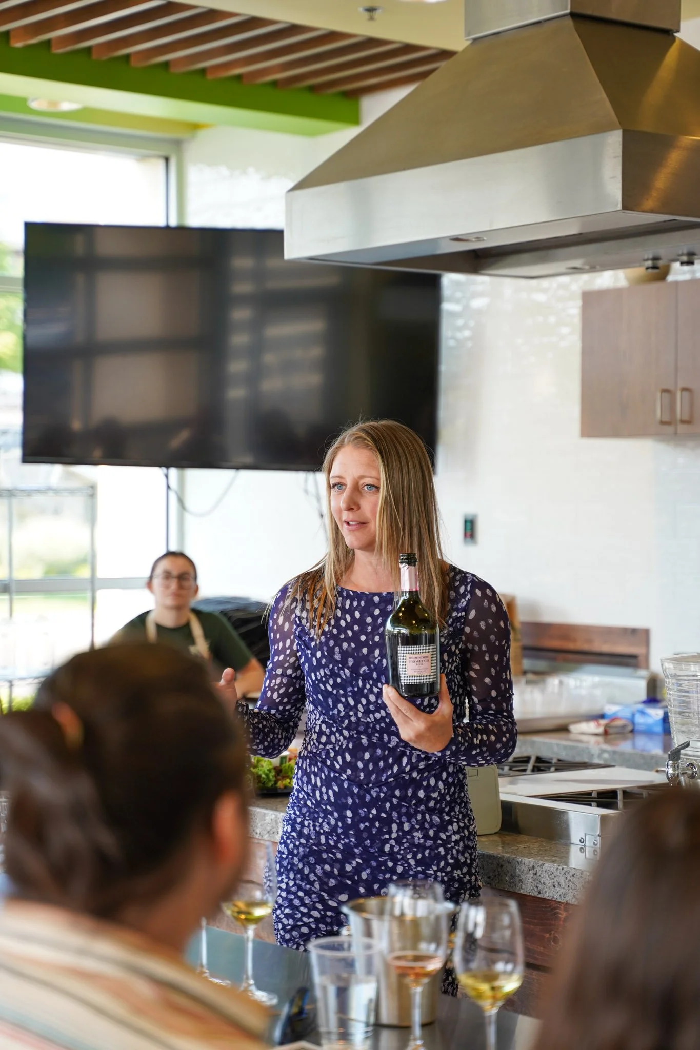 A woman with long blonde hair holding a wine bottle and speaking to a group in a modern kitchen or restaurant setting, with other people seated around the table. There are wine glasses on the table and a large flat-screen TV on the wall.