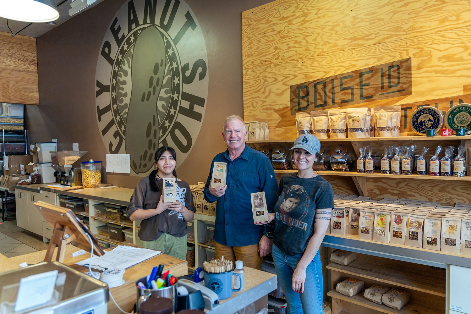 Three people inside a store, smiling at the camera, holding packages of snacks. The store has shelves with packaged snacks and a large logo on the wall with a pickle and the words 'PEANUT BUTTER'.