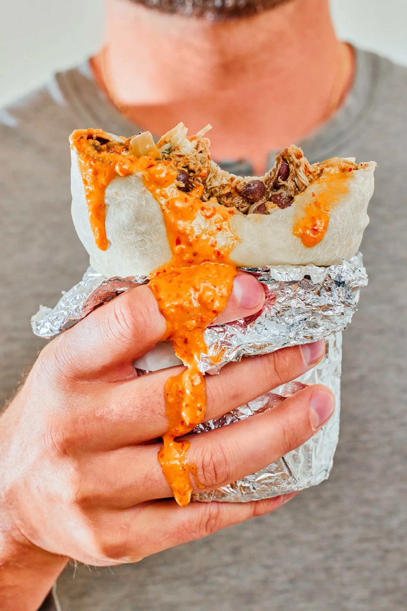 Close-up of a hand holding a burrito wrapped in aluminum foil, with melted cheese and shredded meat spilling out, against a blurred background of a person in a gray shirt.