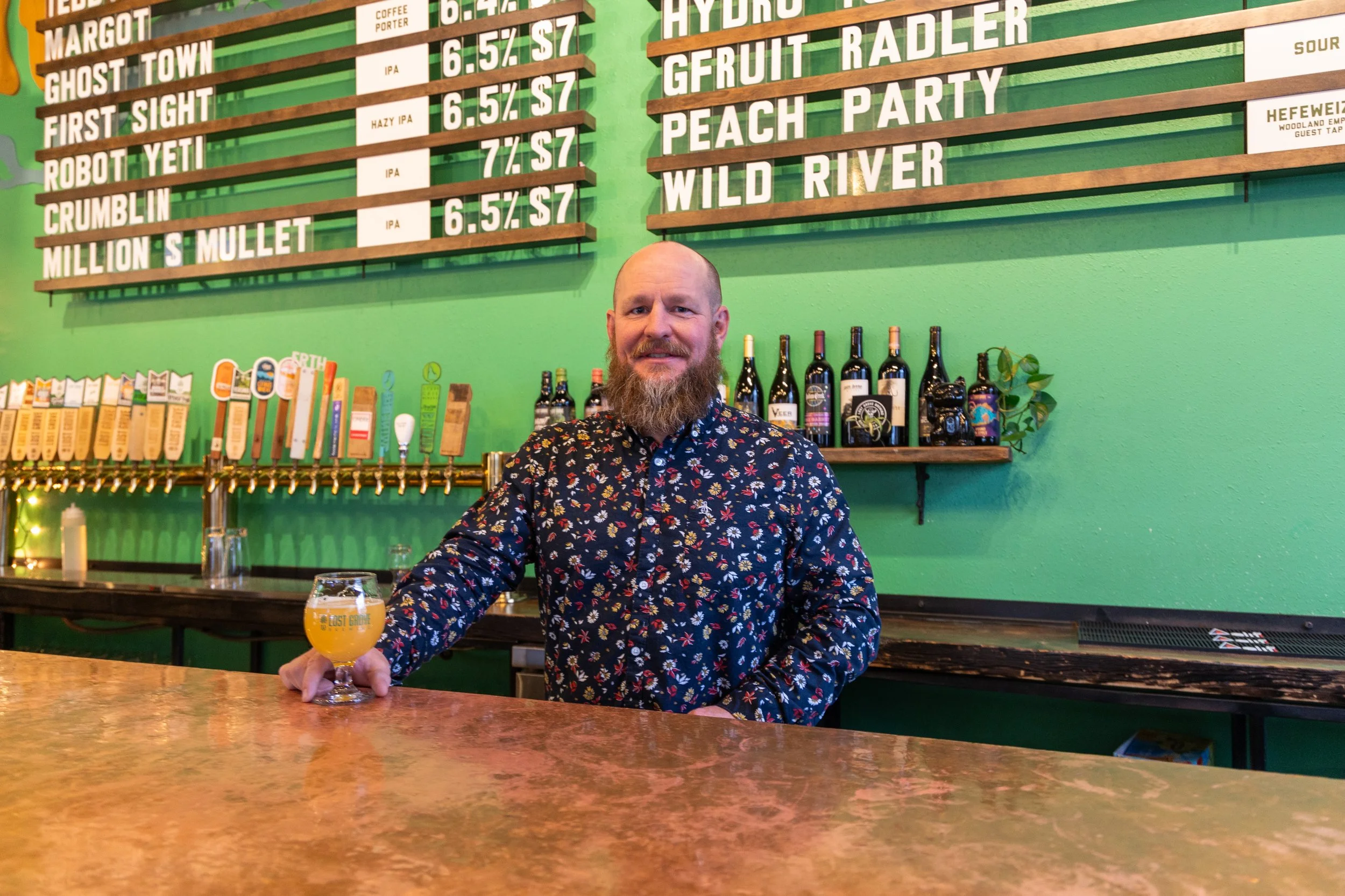 A man with a beard wearing a dark floral shirt sitting at a bar counter with a glass of beer, in front of a green wall with a large beer menu and bottles behind him.