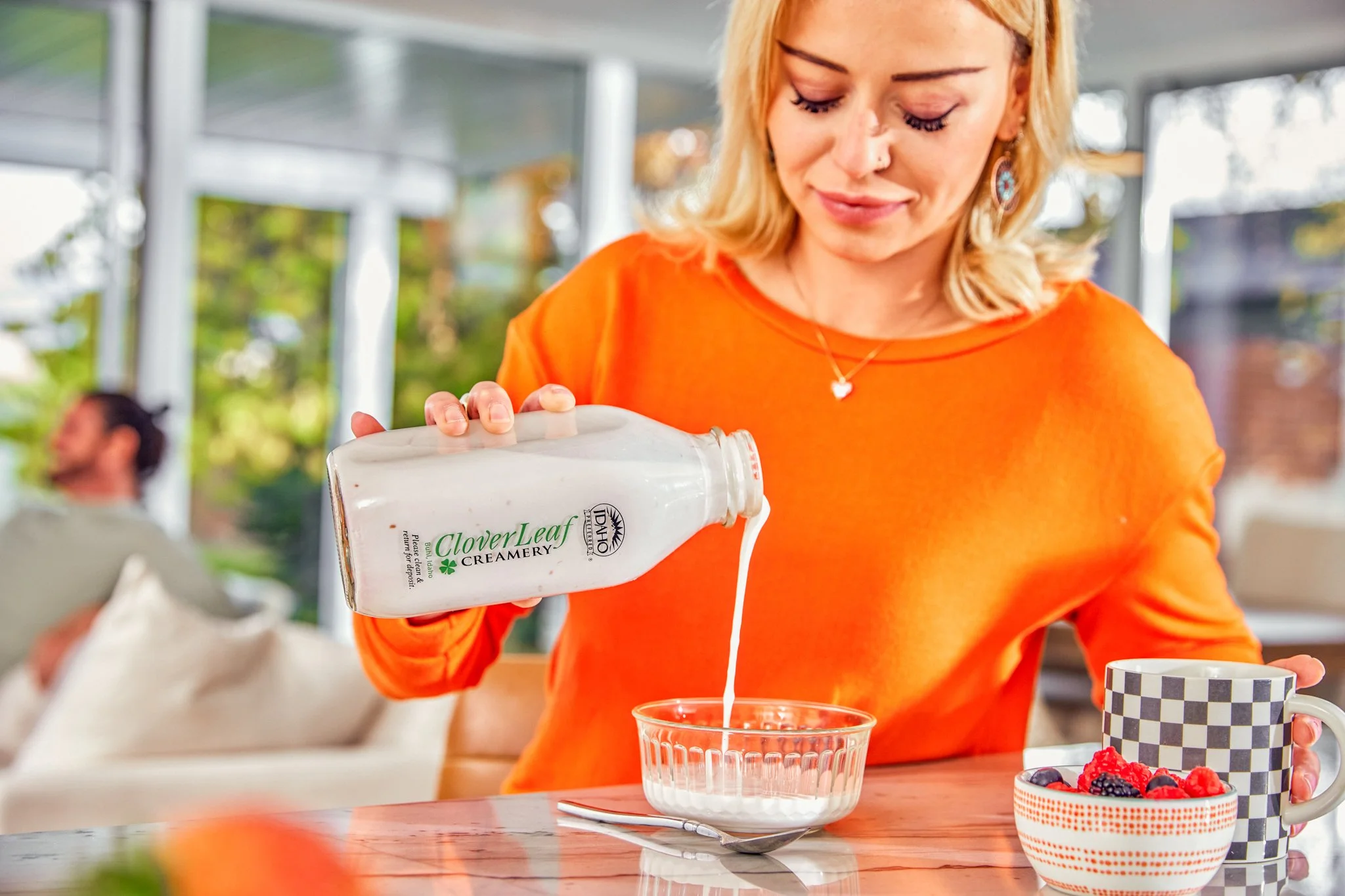 Woman with blonde hair pouring cream from Clover Leaf Creamery container into a glass on a kitchen table, with a checkered mug and berries nearby.