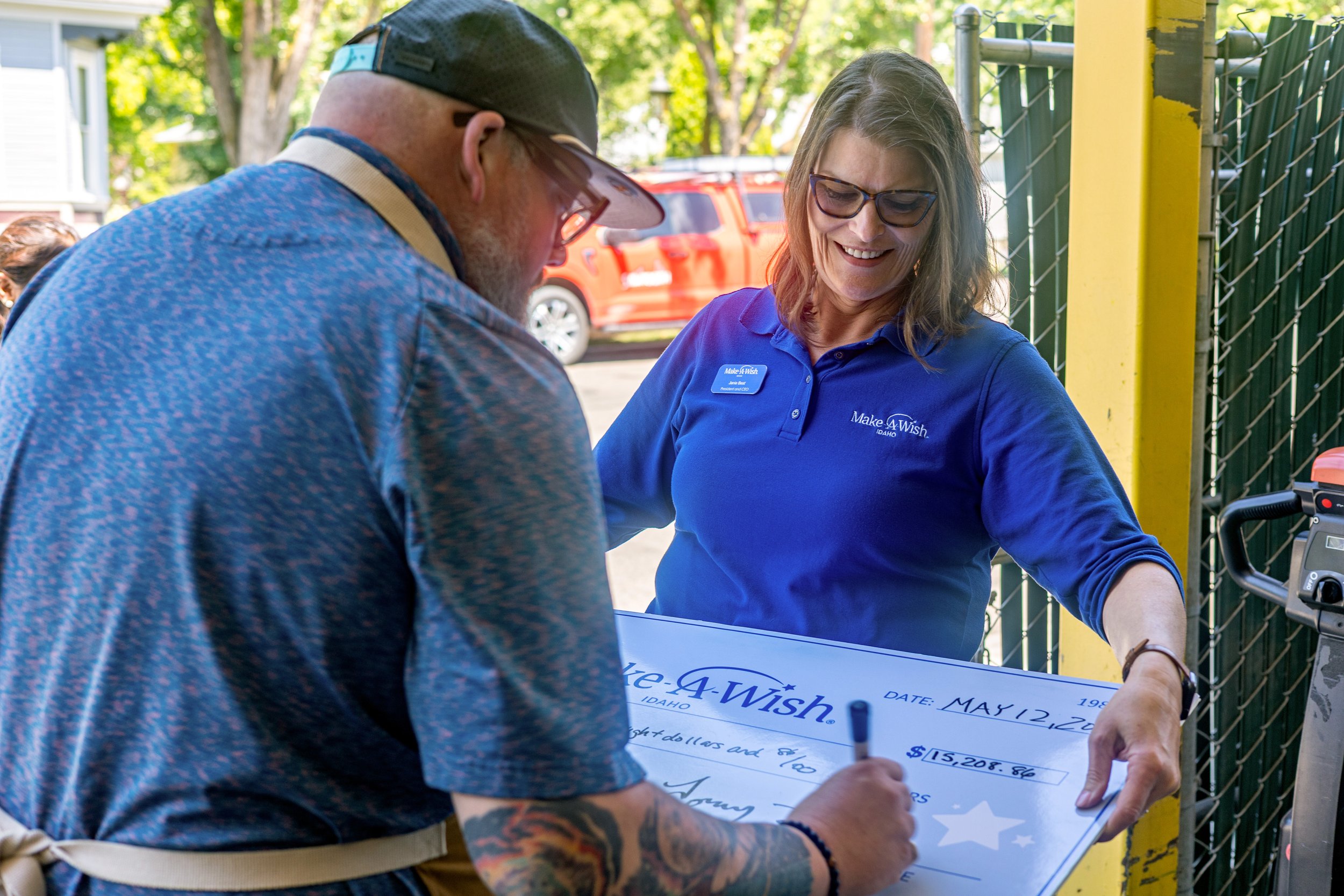A man with tattoos on his arm signs a giant check at a Make-A-Wish event, while a woman in a blue Make-A-Wish shirt observes. They are outdoors near a fence with trees and a red vehicle in the background.