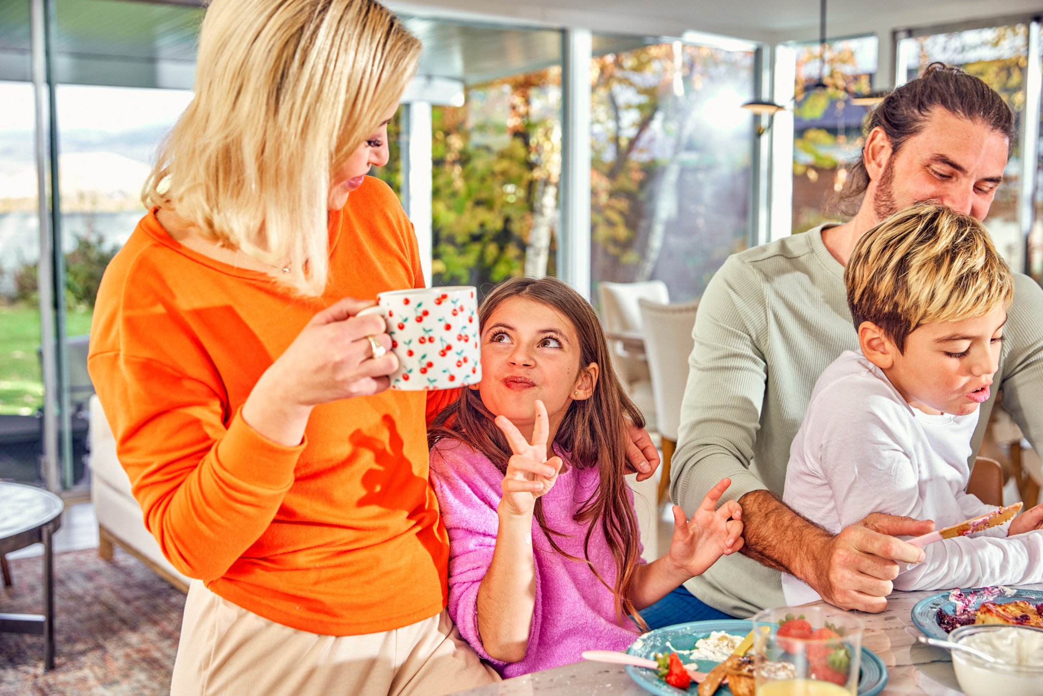 A woman in an orange top holding a cup, looking at a girl in a pink top making a peace sign, while a man and a boy sit nearby during a meal in a bright indoor space with large windows and trees outside.