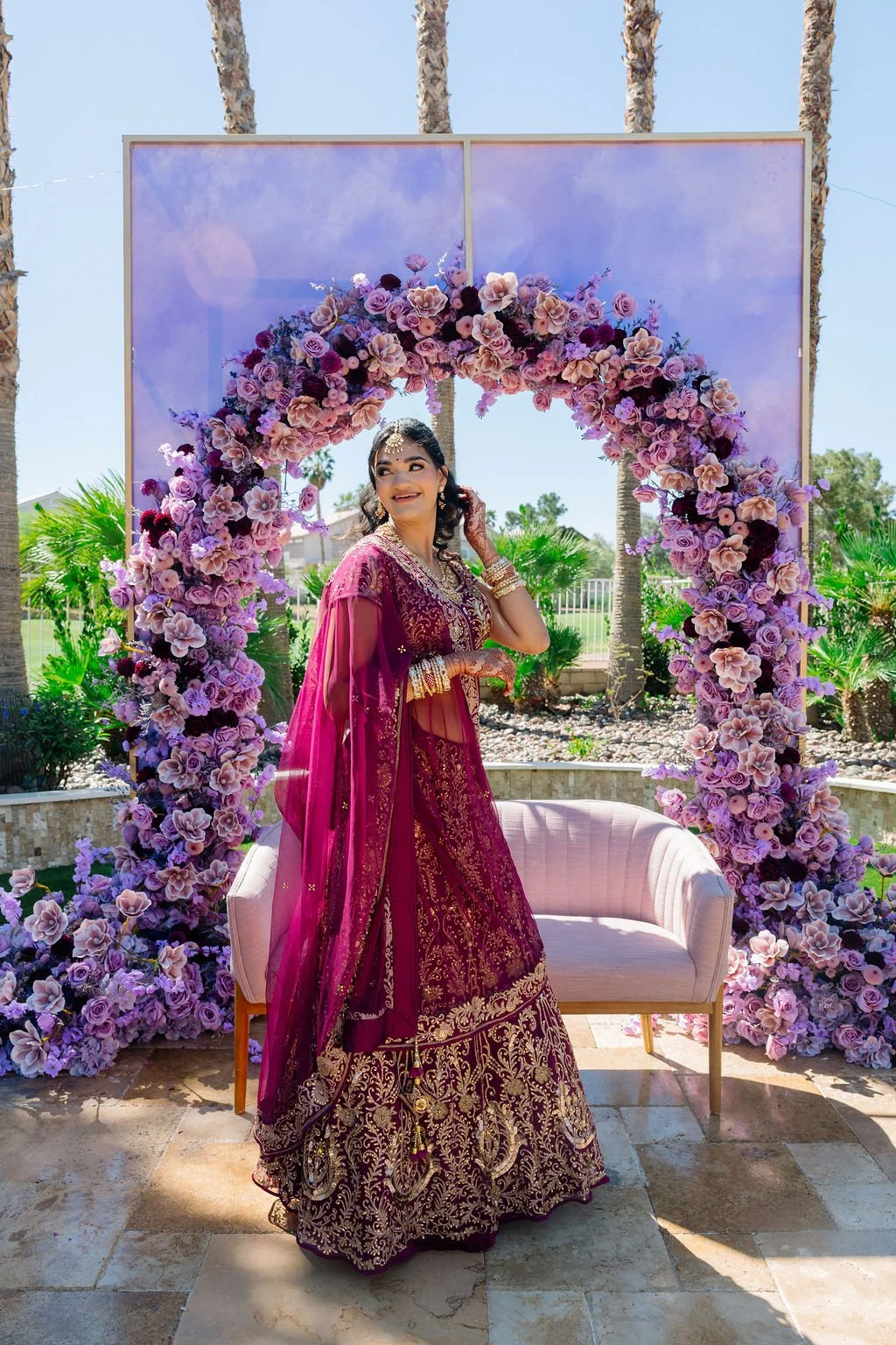 A woman dressed in a maroon and gold traditional Indian outfit, standing under a purple floral arch at an outdoor event, smiling and posing for the photo.