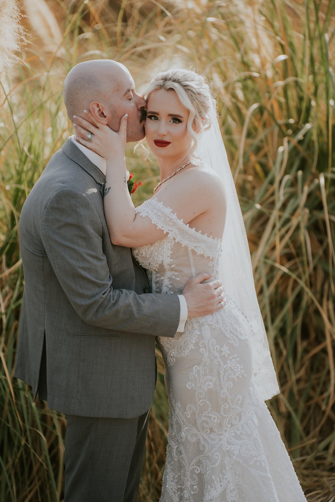 A bride and groom in wedding attire sharing an intimate moment outdoors, surrounded by tall grass.