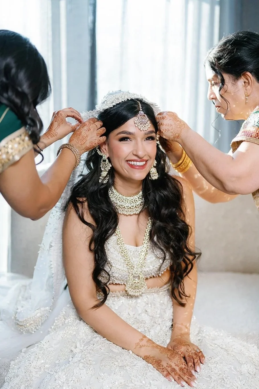 Bride in traditional wedding attire being adorned with jewelry by two women, sitting on a sofa with a smile.