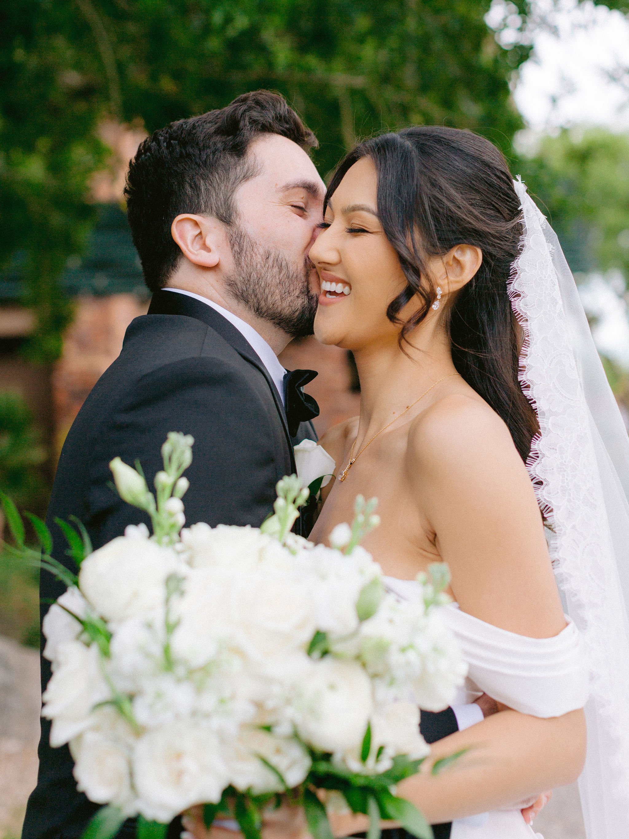 Close couple portrait with groom kissing bride's cheek as she laughs at Las Vegas Paiute Golf Resort wedding, hair by Kari K and makeup by Natalia W for Amelia C & Co, photography by Susie and Will