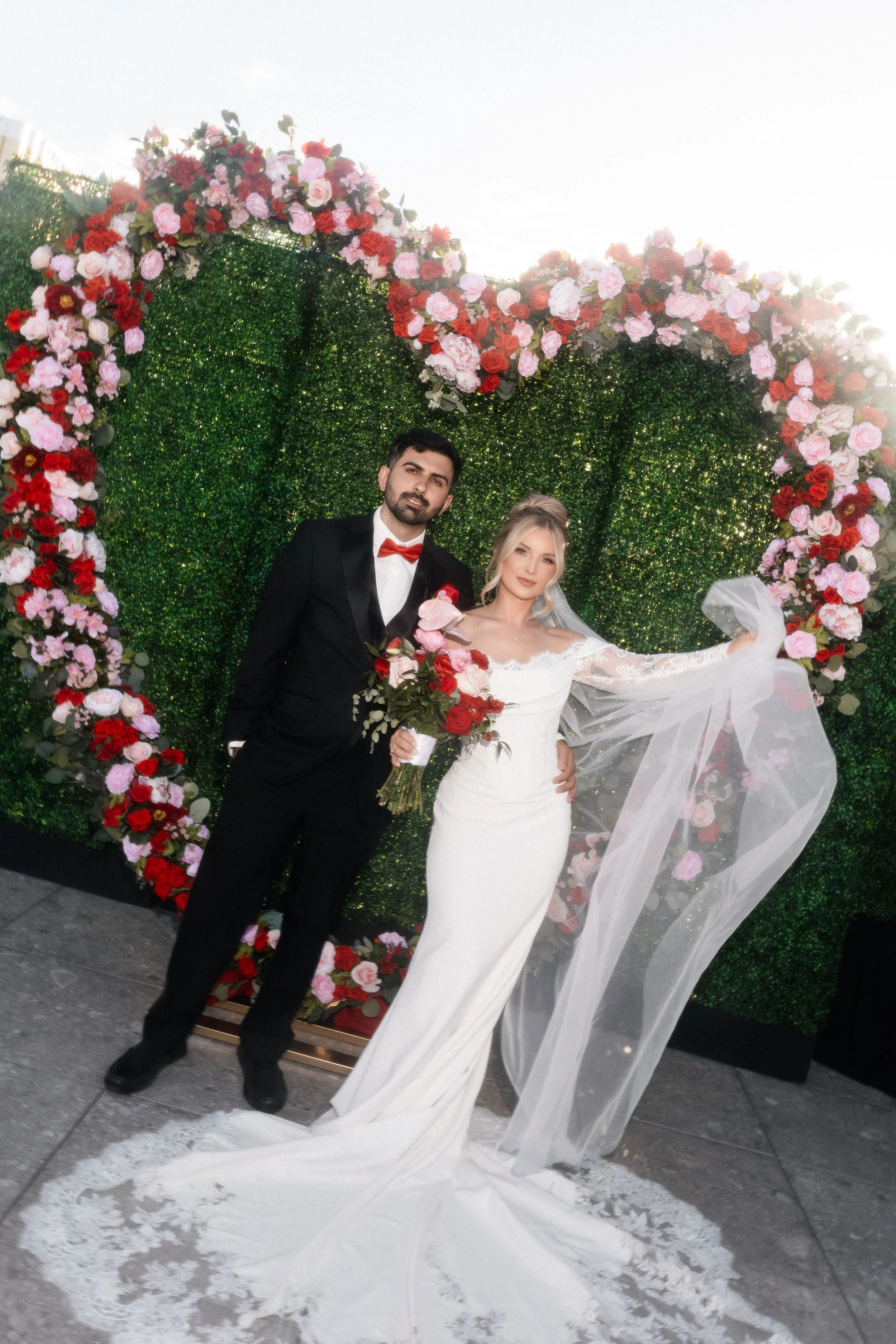 Bride and groom posing in front of floral heart arch with veil flowing at Las Vegas wedding, hair by Katie A and makeup by Aimee A for Amelia C & Co, photography by Risky Exposure