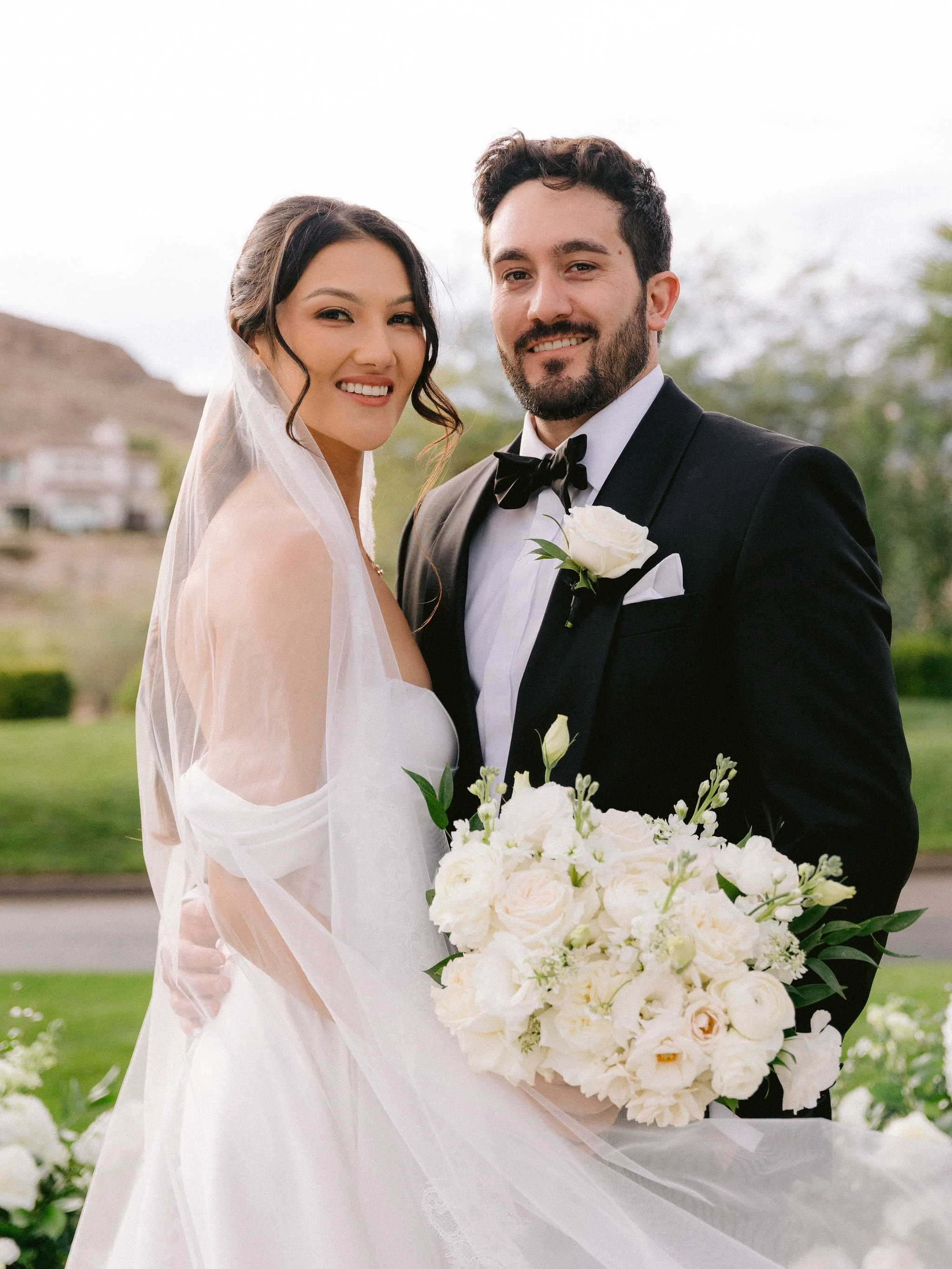 Bride and groom smiling at camera at Las Vegas Paiute Golf Resort wedding with golf course backdrop and white bouquet, hair by Kari K and makeup by Natalia W for Amelia C & Co, photography by Susie and Will