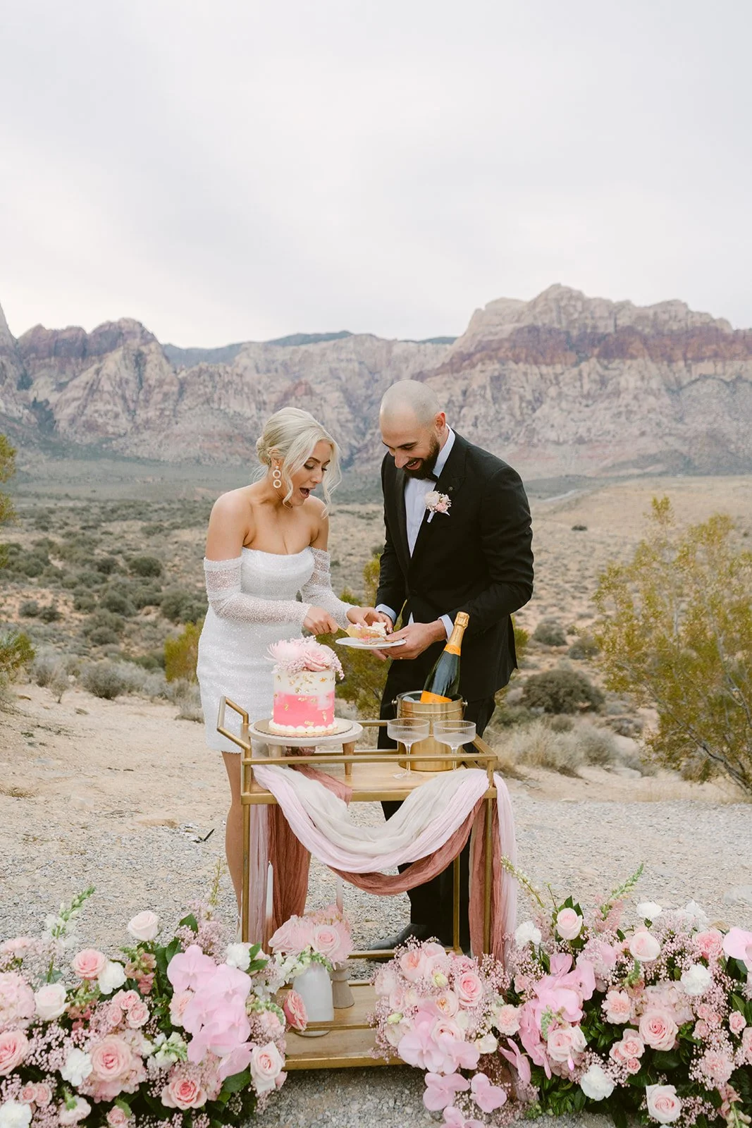 Bride and groom cutting pink wedding cake on gold bar cart at Red Rock Canyon Las Vegas elopement surrounded by pink florals, hair and makeup by Luci L for Amelia C & Co, photography by Kristen Joy