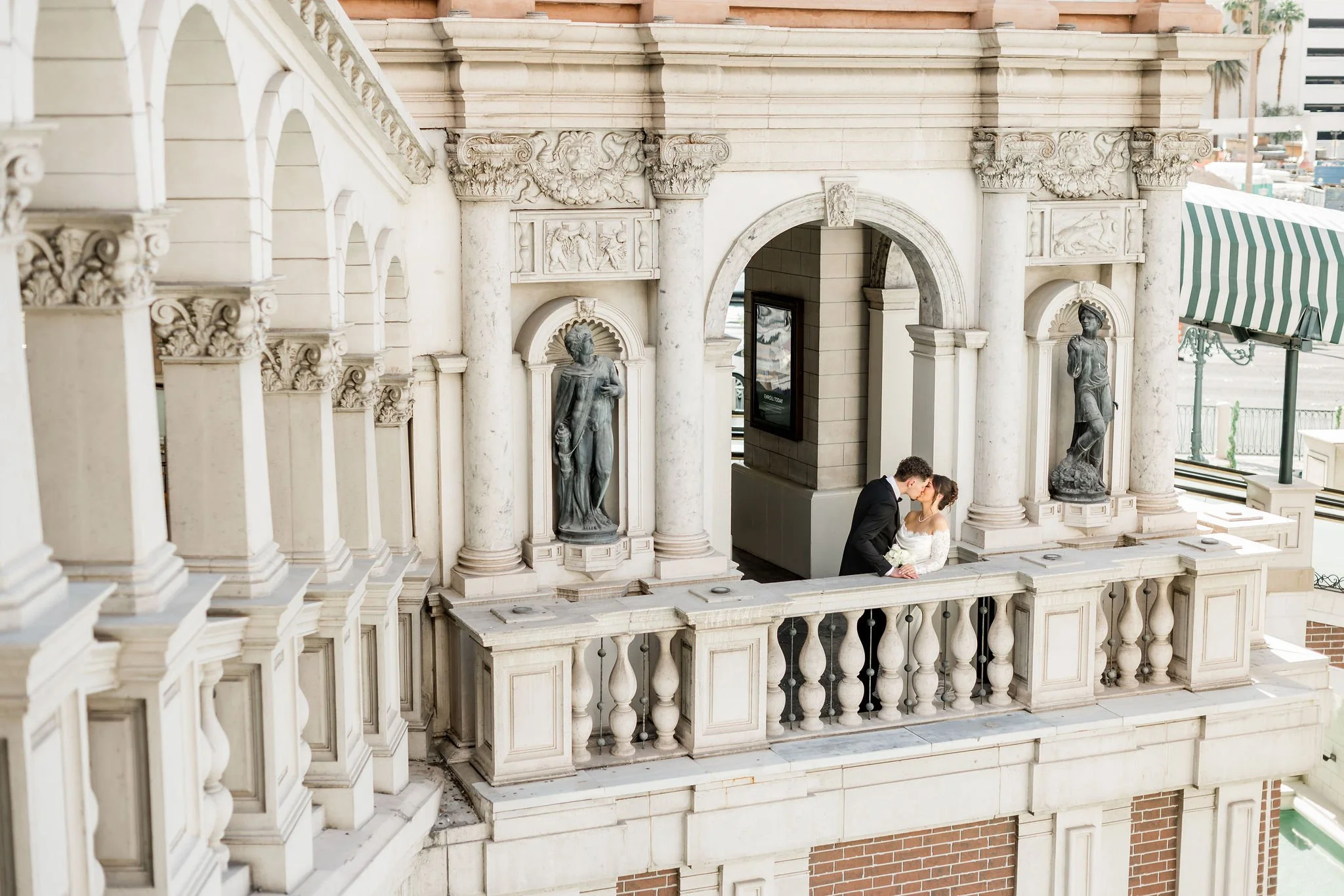 Wide architectural shot of bride Jessica and groom Lukas kissing on The Venetian Las Vegas balcony with ornate statues, hair by Katie and makeup by Aimee for Amelia C & Co, photography by Liz Le