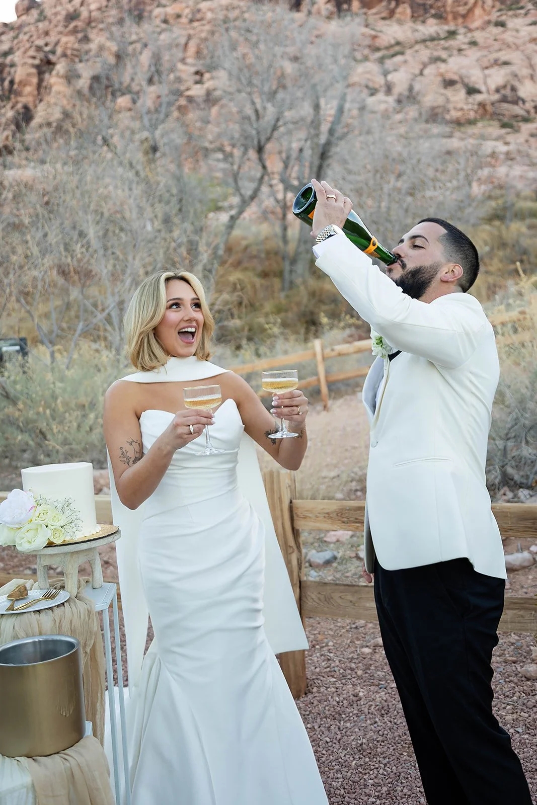 Melysa laughing as Carlos drinks from champagne bottle beside wedding cake at Red Spring Boardwalk Las Vegas elopement, hair by Luci and makeup by Natalia for Amelia C & Co, photography by Onyx & Arrow
