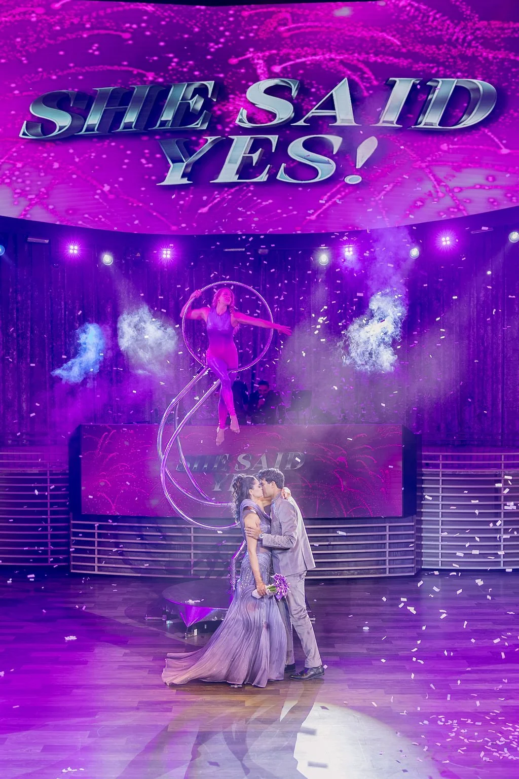 Indian couple kissing on stage during surprise proposal with confetti and aerial performer at Las Vegas engagement celebration, hair by Karen K and makeup by Natalia W for Amelia C & Co, photography by Elizabeth Burgi