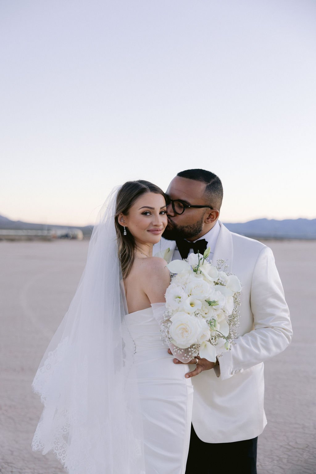 Bruce kissing Cameron's cheek as she smiles at camera at Dry Lake Bed Las Vegas elopement at sunset, hair and makeup by Marai for Amelia C & Co, photography by Sofia Teresa