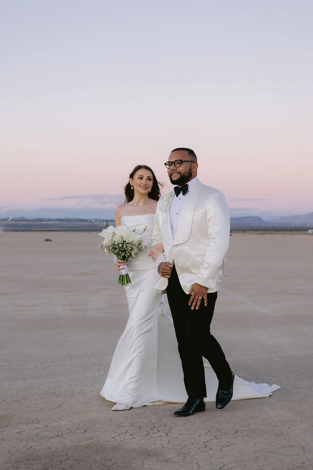 Cameron and Bruce walking together at Dry Lake Bed Las Vegas elopement under pink and purple sunset sky, bride in satin sheath gown with train and groom in white tuxedo, hair and makeup by Marai for Amelia C & Co, photography by Sofia Teresa