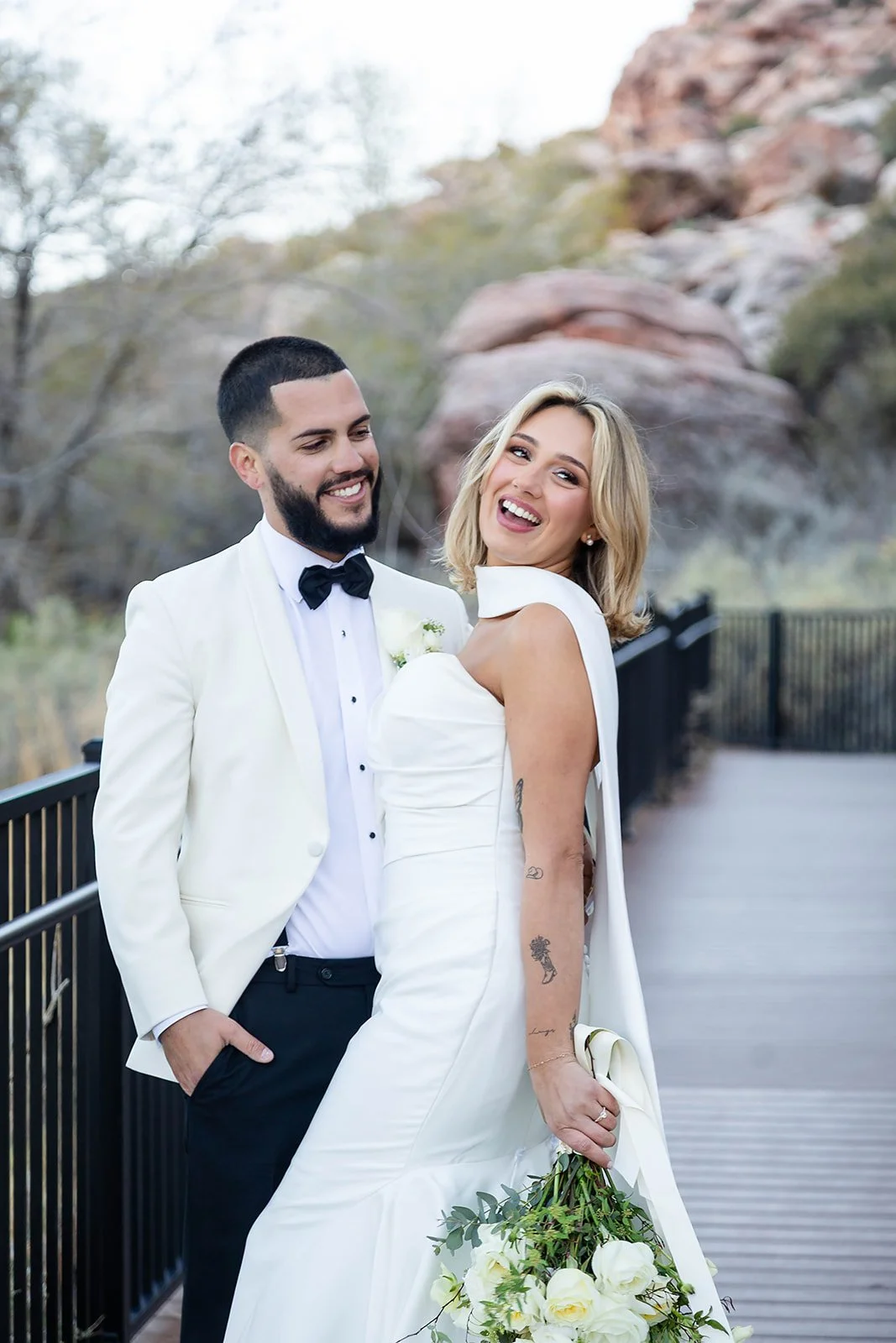 Melysa and Carlos laughing together on bridge at Red Spring Boardwalk Las Vegas elopement, bride in cape halter gown and groom in white tuxedo, hair by Luci and makeup by Natalia for Amelia C & Co, photography by Onyx & Arrow