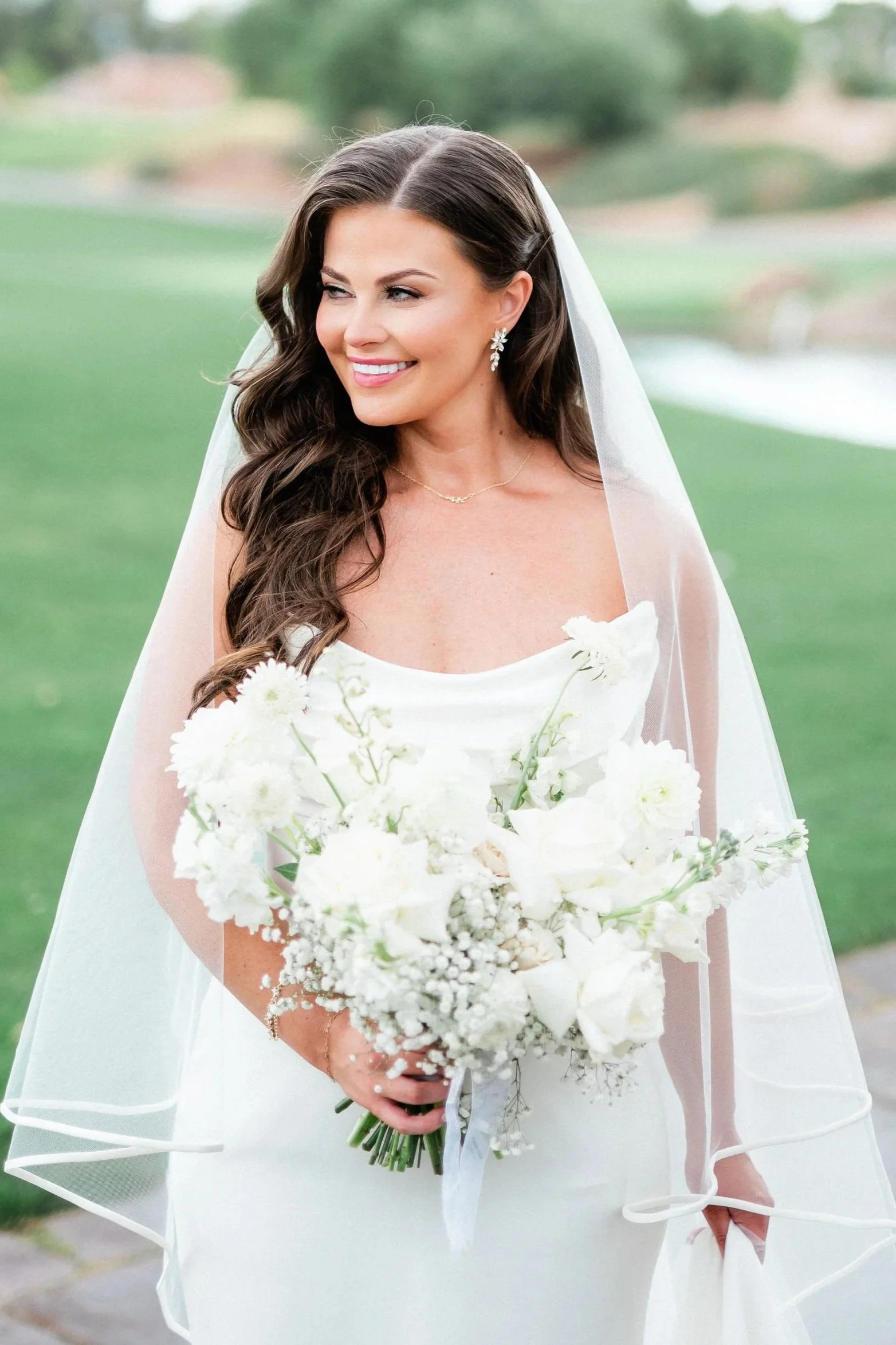 Brunette woman in a white wedding dress holding a bouquet of white flowers, outdoors with greenery in the background.