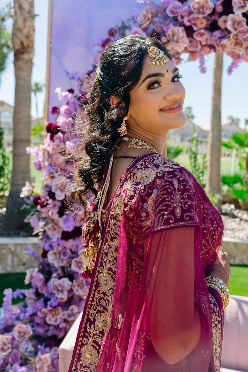  Indian bride in magenta embroidered lehenga standing under purple floral arch at Las Vegas engagement celebration, hair by Karen K and makeup by Natalia W for Amelia C & Co, photography by Elizabeth Burgi, planning by Scheme Events