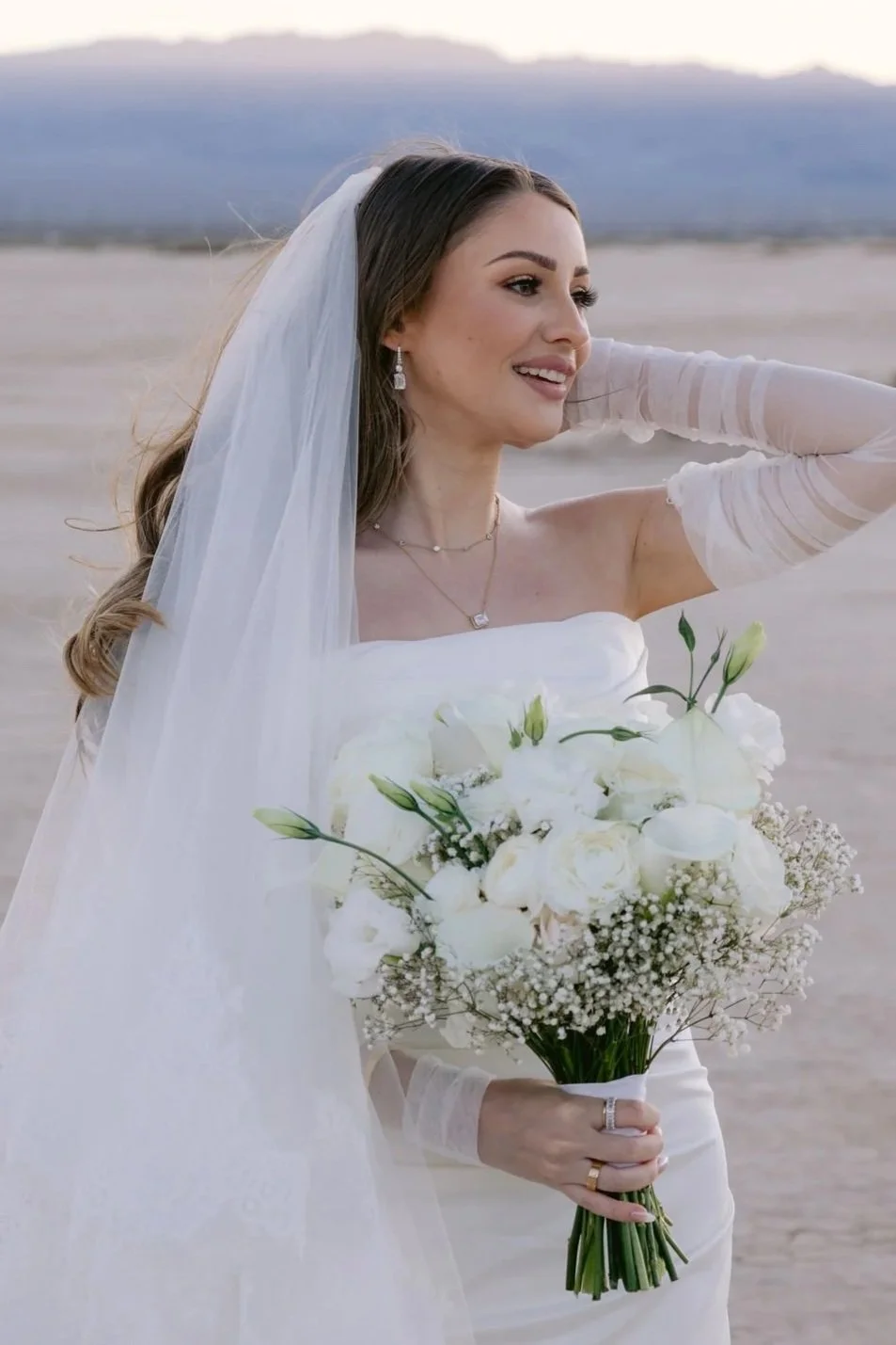 A bride standing on a sandy desert landscape, holding a bouquet of white flowers, wearing a white wedding gown, veil, and jewelry, with mountains in the background.