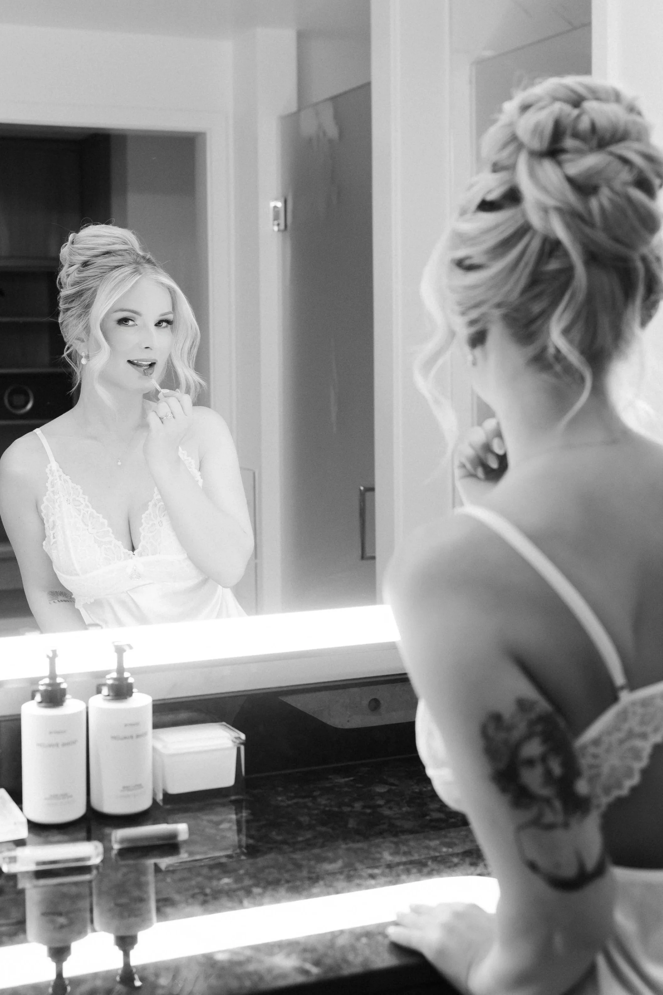 A woman is applying lipstick while looking at herself in the mirror, with her reflection showing her face and upper body, wearing a white lace camisole and jewelry, in a bathroom or dressing area with bottles and toiletries on the counter.