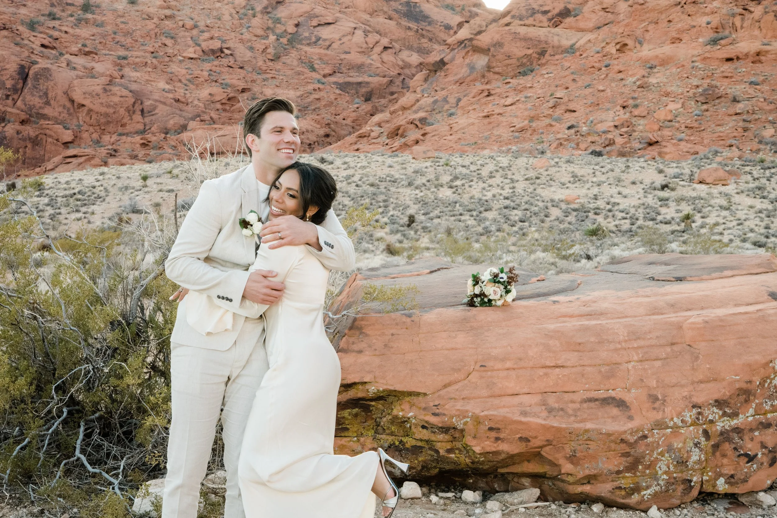 Martinique and James embracing with red rock formation and bouquet at Red Rock Canyon elopement, hair and makeup by Luci for Amelia C & Co, photography by Cactus Collective Weddings
