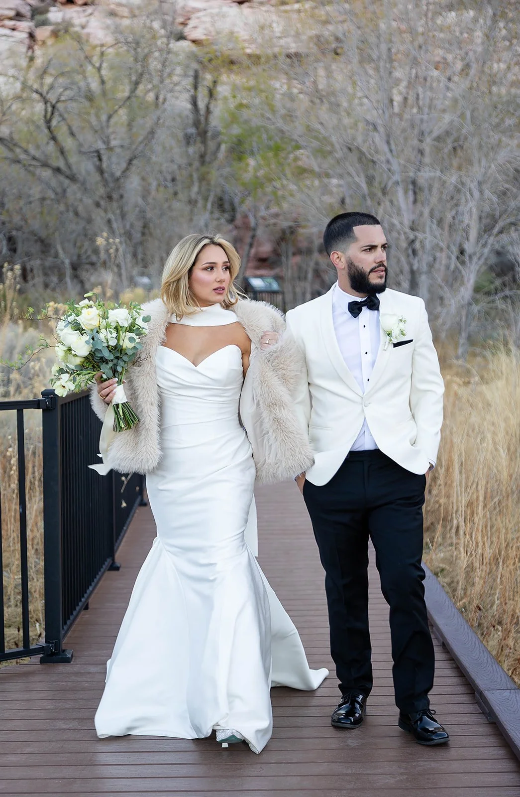 Full-length portrait of Melysa and Carlos on bridge at Red Spring Boardwalk Las Vegas elopement, bride's train spread and red rock formations behind, hair by Luci and makeup by Natalia for Amelia C & Co, photography by Onyx & Arrow