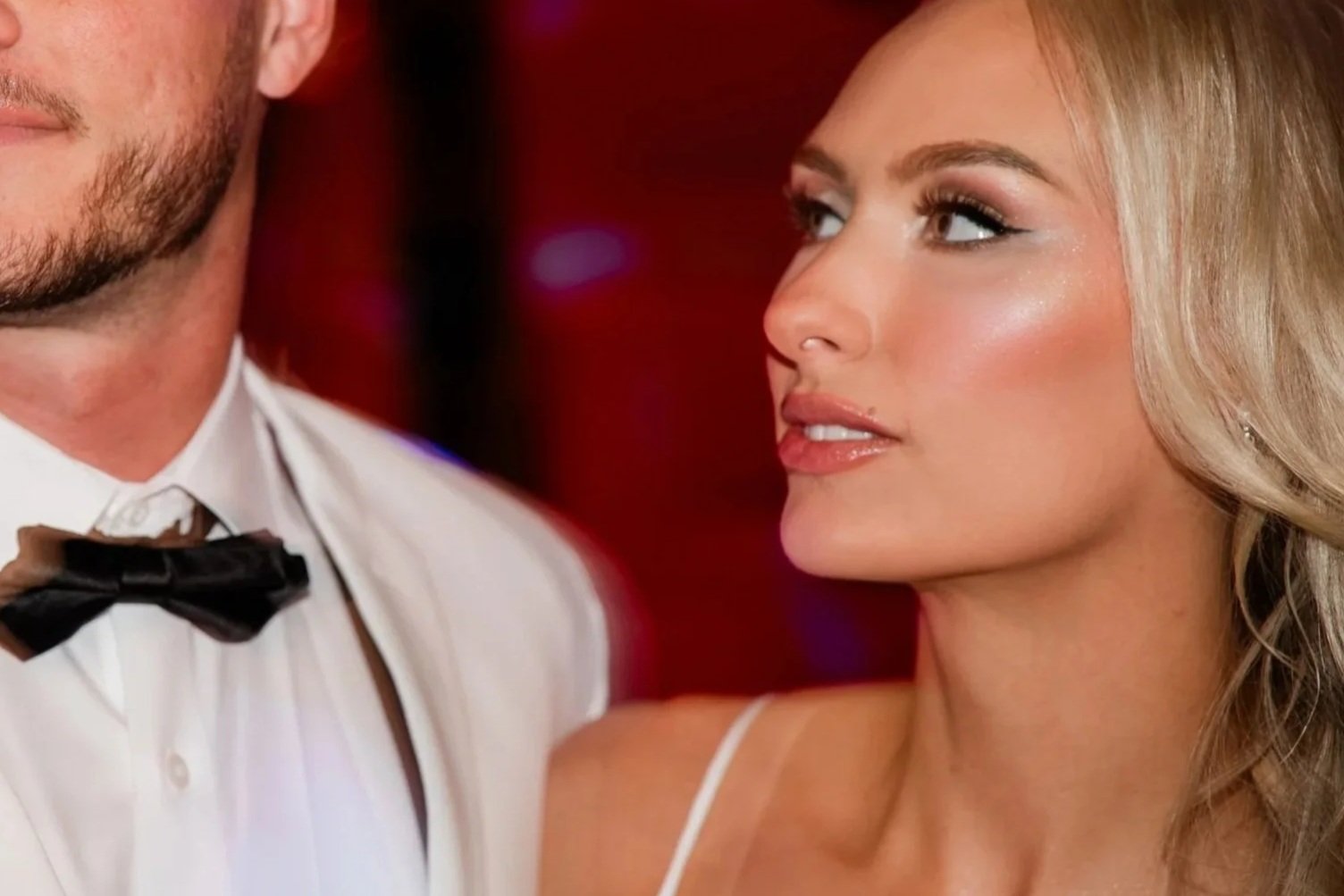 Close-up of a woman with blonde hair, makeup, and a white strap dress looking at a man with a beard in a tuxedo with a bow tie, against a red background.