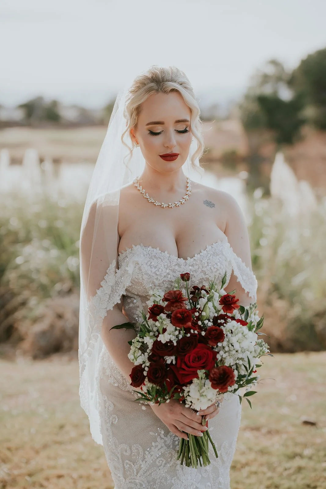 A bride with blonde hair in an updo, wearing a strapless lace wedding dress, holding a bouquet of red and white flowers, standing outdoors with a blurred background of trees and grass.