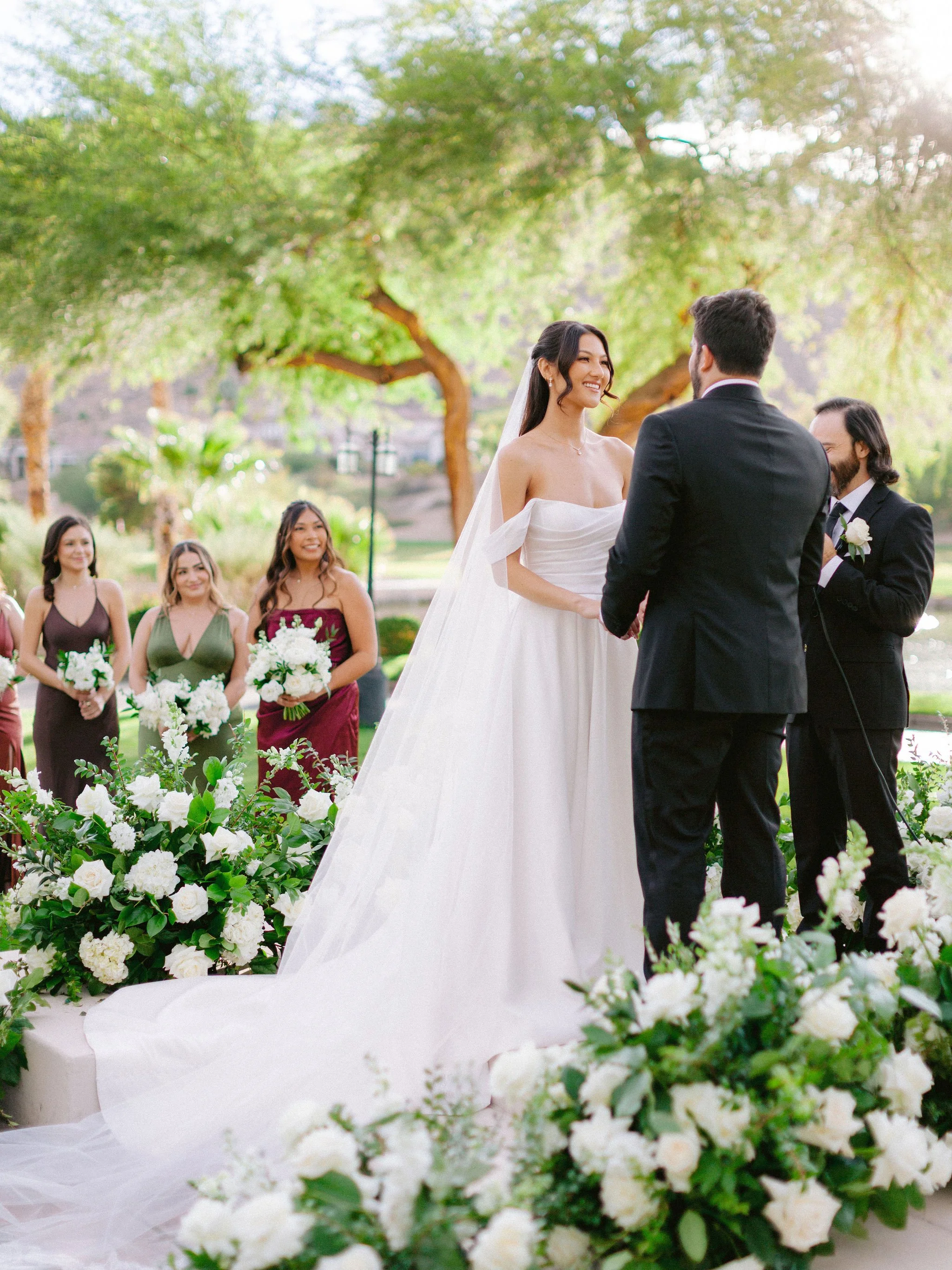 Outdoor wedding ceremony at Las Vegas Paiute Golf Resort with bride smiling at groom surrounded by white floral arrangements and bridesmaids in jewel tone dresses, hair by Kari K and makeup by Natalia W for Amelia C & Co, photography by Susie and Wil