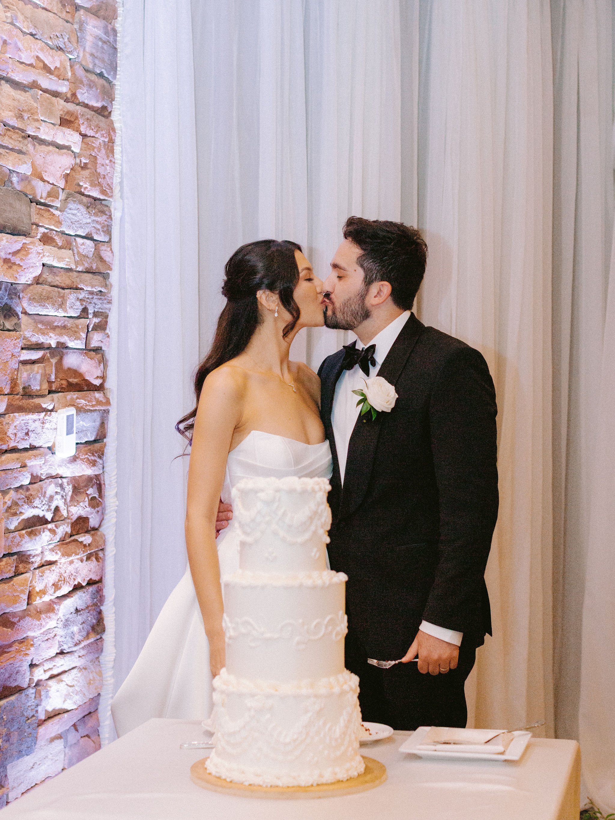 Bride and groom kissing during cake cutting at Las Vegas Paiute Golf Resort wedding reception, bride with second look half updo, hair by Kari K and makeup by Natalia W for Amelia C & Co, photography by Susie and Will