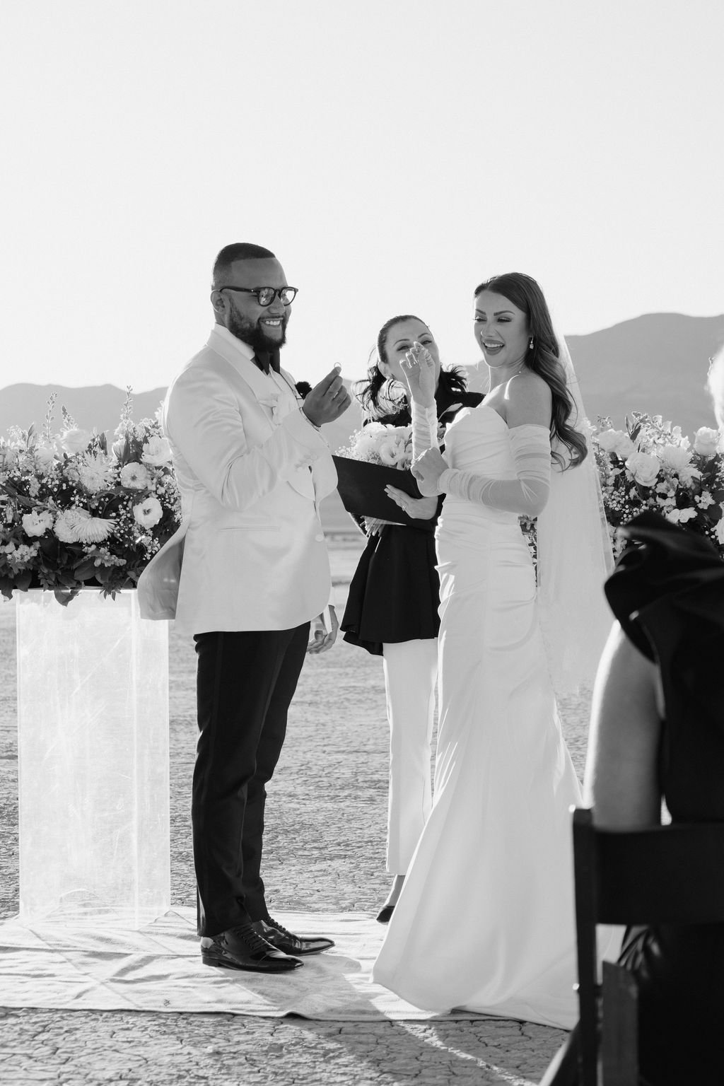Cameron and Bruce laughing during ceremony at Dry Lake Bed Las Vegas elopement with white floral altar arrangements, hair and makeup by Marai for Amelia C & Co, photography by Sofia Teresa