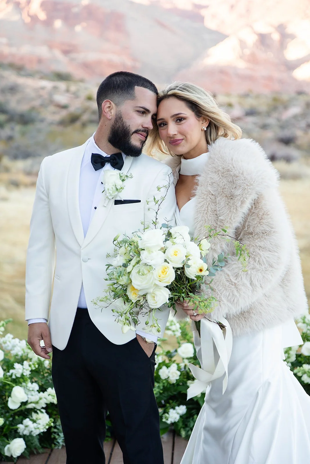 Melysa and Carlos on bridge at Red Spring Boardwalk Las Vegas elopement, bride in fur stole over cape gown and groom in white tuxedo, hair by Luci and makeup by Natalia for Amelia C & Co, photography by Onyx & Arrow