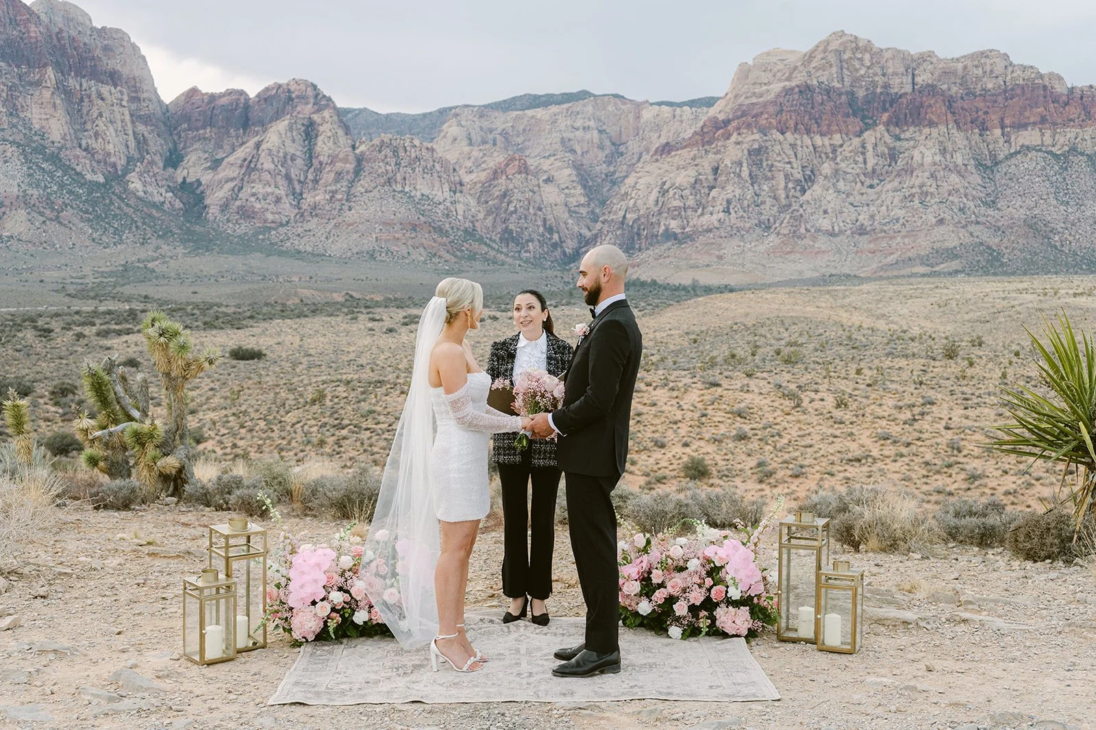 Bride and groom exchanging vows at Red Rock Canyon Las Vegas elopement with pink floral arrangements and desert backdrop, hair and makeup by Luci L for Amelia C & Co, photography by Kristen Joy