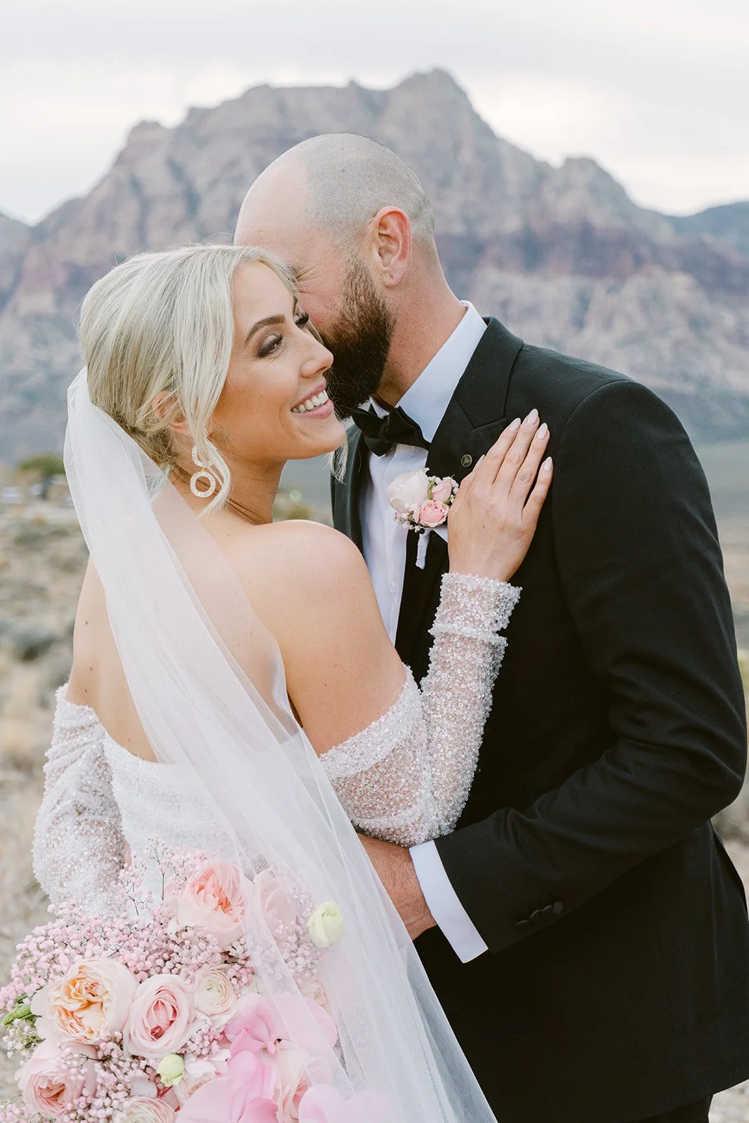 Intimate couple portrait at Red Rock Canyon Las Vegas elopement, groom kissing bride's temple with desert mountain backdrop, hair and makeup by Luci L for Amelia C & Co, photography by Kristen Joy